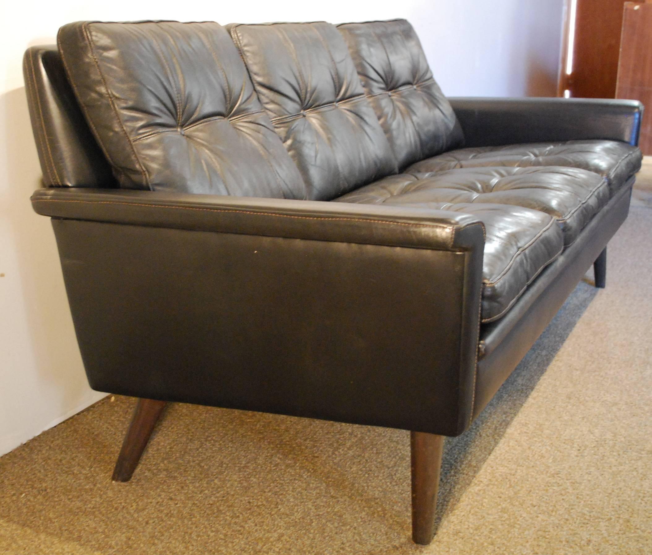 A Hans Olsen designed Danish Mid-Century Modern three-cushion sofa in black full grain leather showing button tufted seats and backrests and graceful curved armrests with contrasting stitching. Note the single hide back panel.