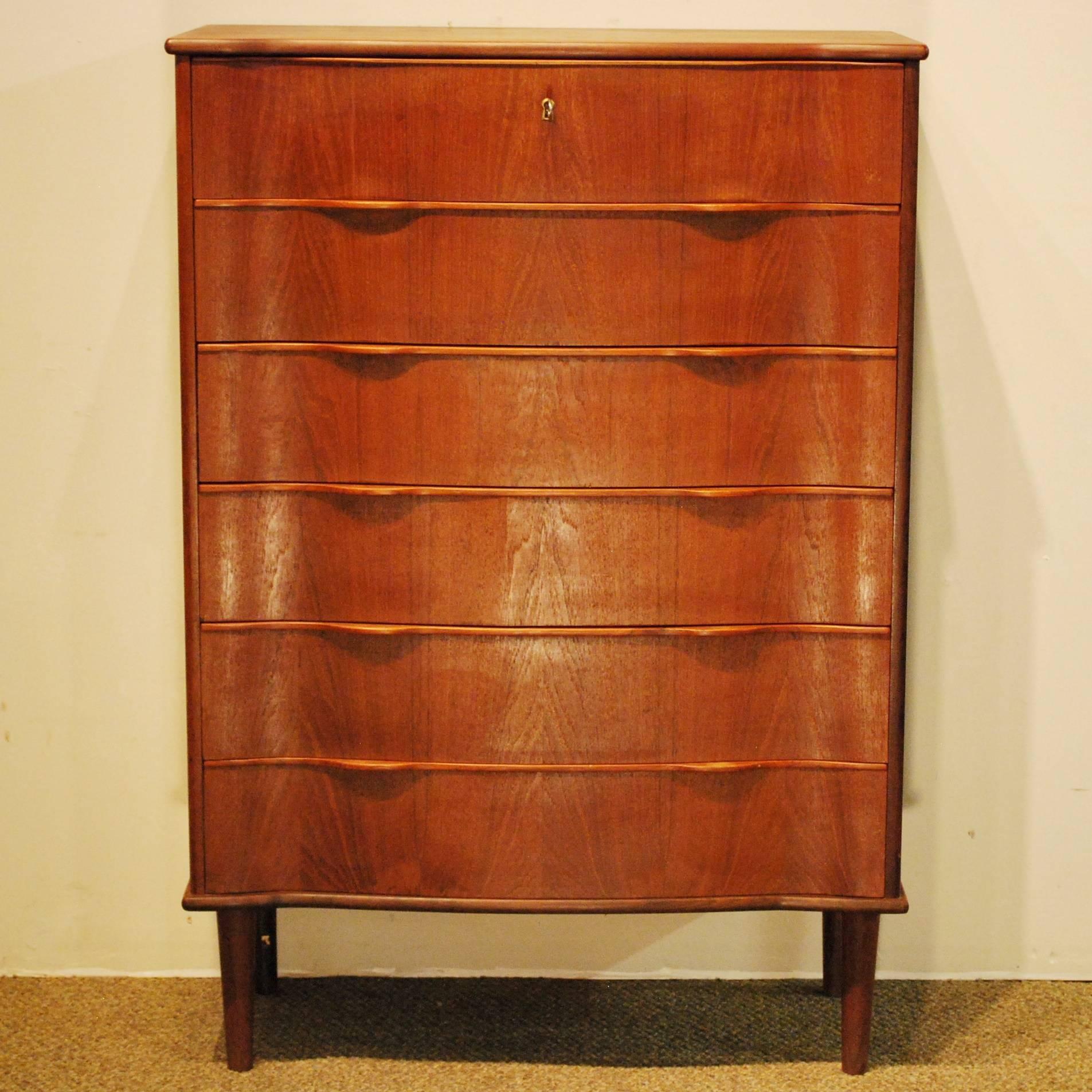 
A Mid-Century Modern serpentine front tall chest featuring six dovetailed drawers with integrated shaped pulls, a locking top drawer with key, softly beveled teak self-banding edges, and beautiful matched grain teak construction.