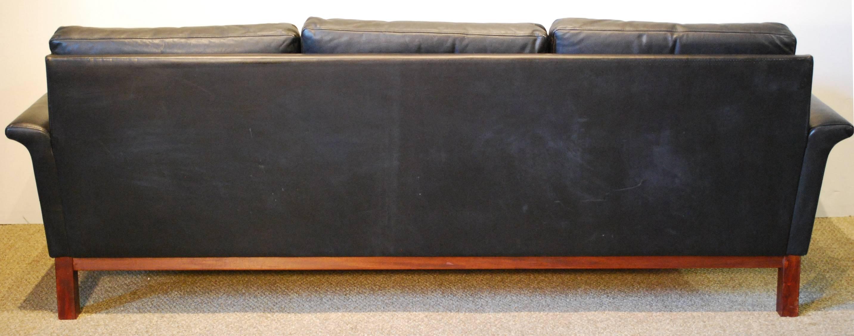 A Hans Hansen designed Danish modern full grain black leather sofa featuring a Classic three cushion design with gracefully rolled armrests and a wooden base.