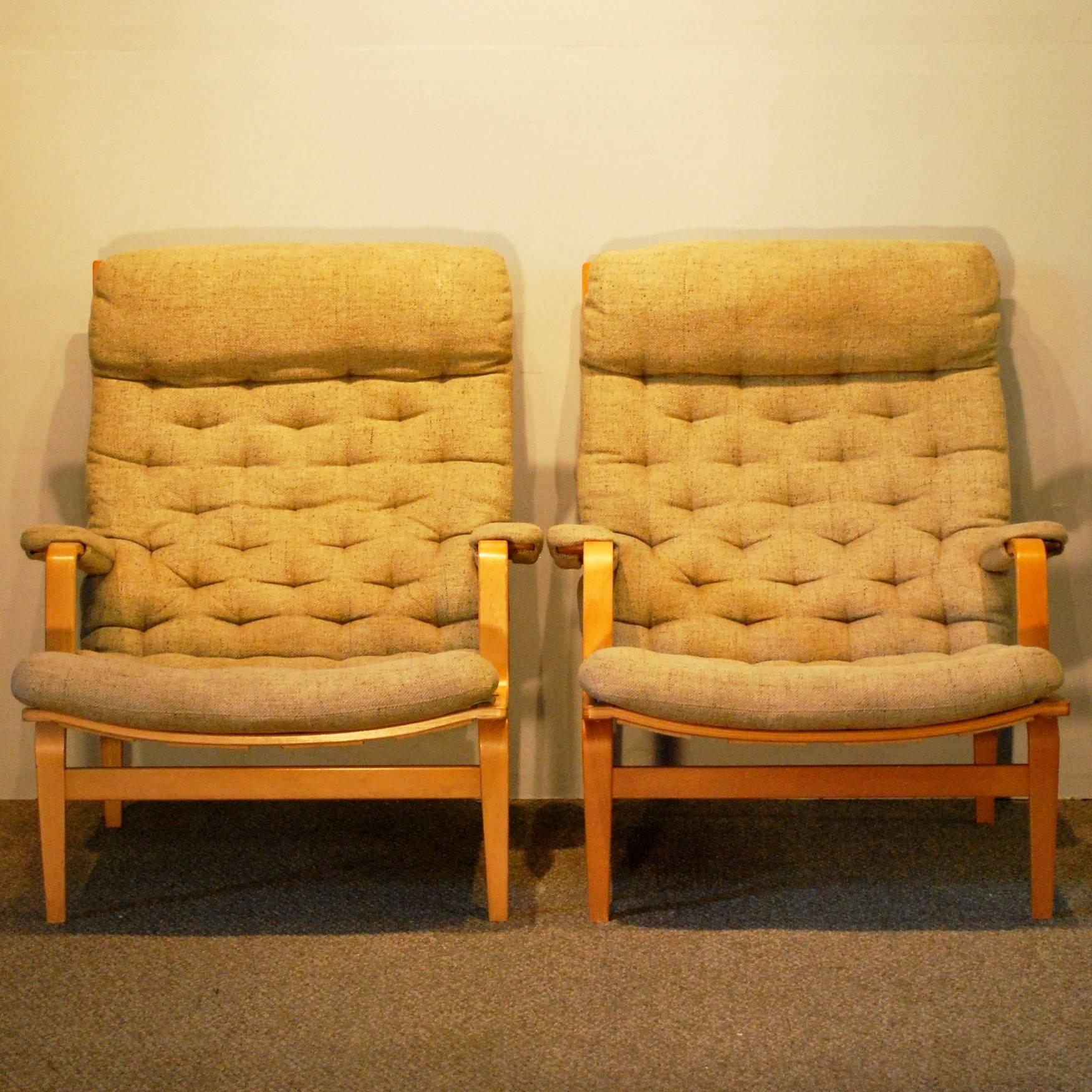 A Bruno Mathsson designed for DUX Mid-Century Modern pair of 'Ingrid' lounge chairs displaying Classic high back bentwood beech frames with button stitched upholstered cushioning and cushioned armrests with a fabric backing.