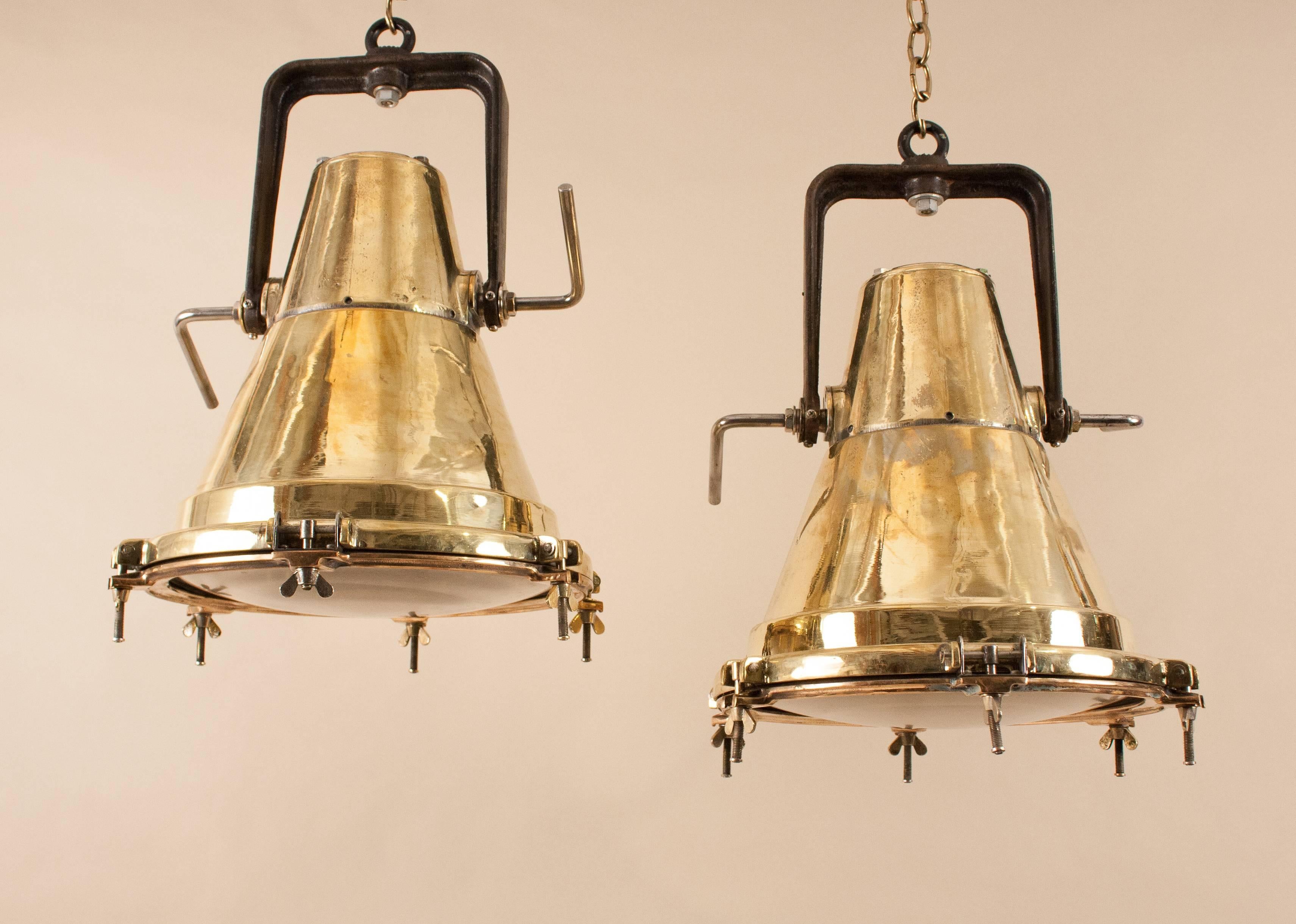An authentic and uncommon pair of 1950s polished brass ship deck lights with wonderful size, shape and patina. Salvaged from a marine vessel and attentively restored, these nautical pendants suspend from adjustable black iron brackets and have