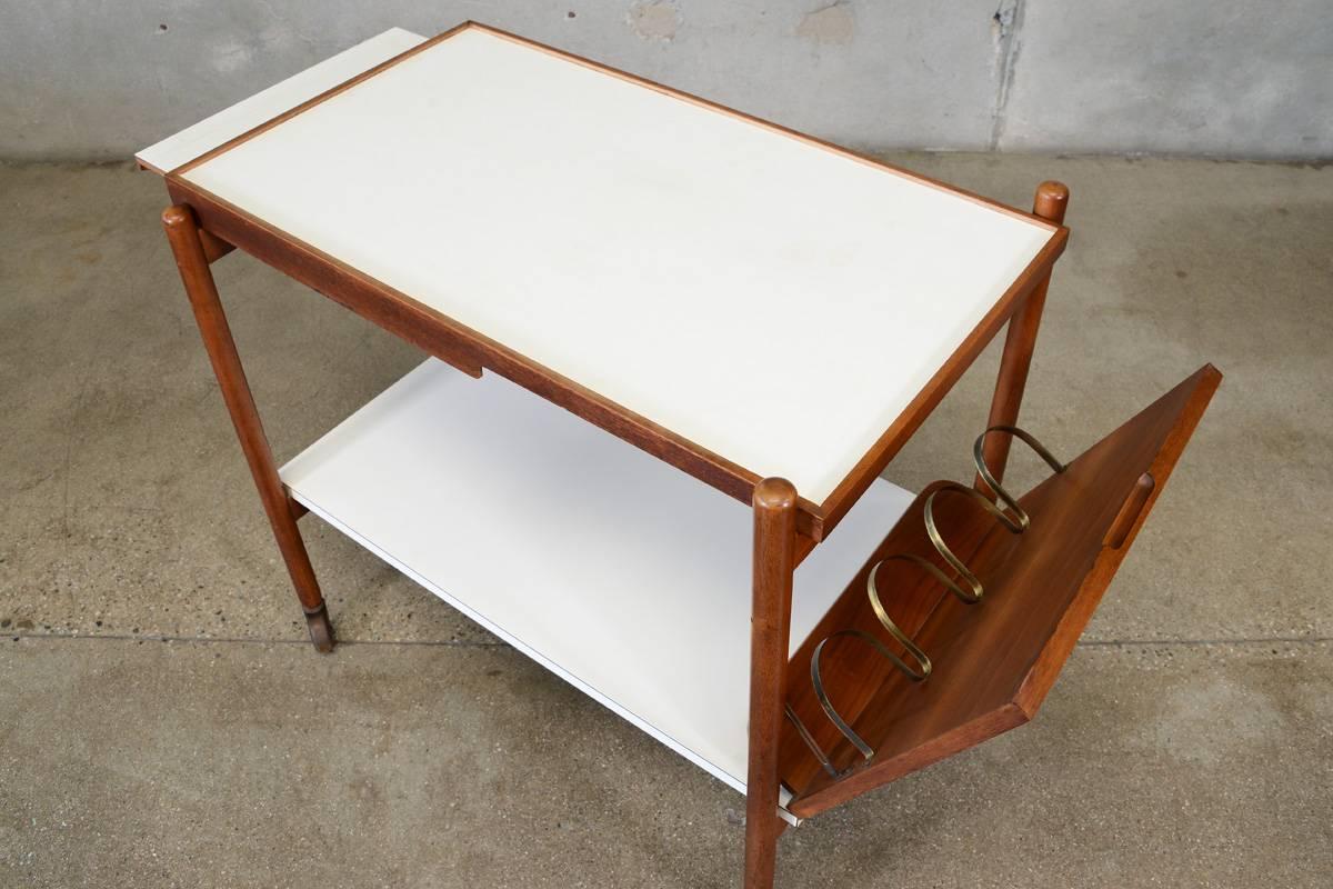 A rarely seen bar cart designed by Greta Grossman for Glenn of California. Walnut construction with white melamine tops and brass accents. The upper level has a pull-out serving tray, while the lower lever offers a unique tilt out bottle holder.
