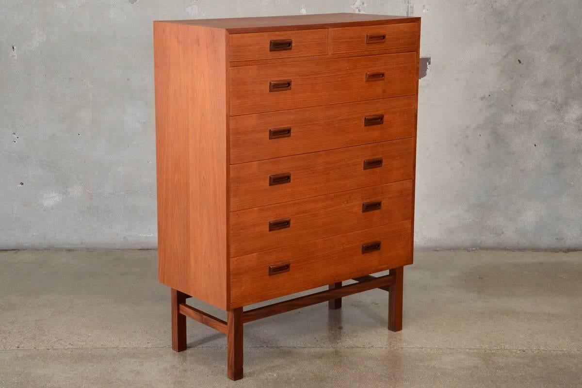 One of a pair we currently have available, this Danish teak high boy dresser offers ample storage with Classic Minimalist styling reminiscent of Borge Mogensen. The five spacious full width drawers and two small top drawers are accent with sculpted