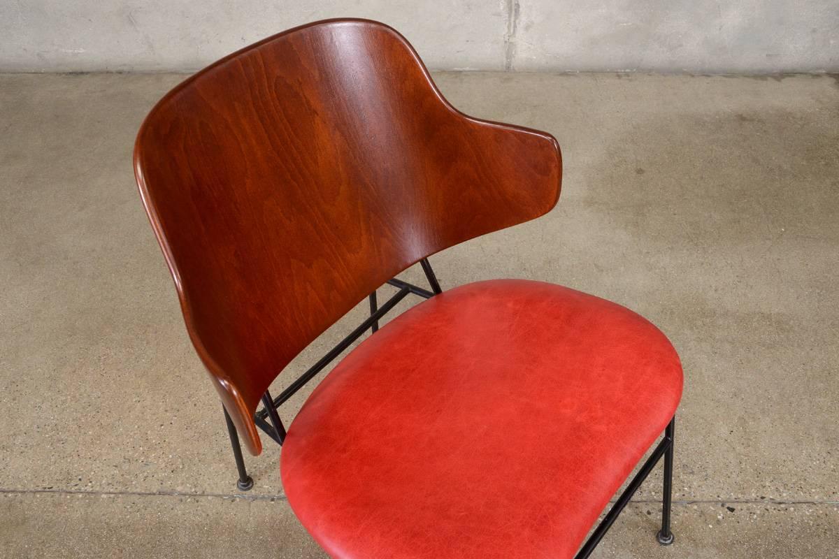 A classic “Penguin” lounge chair designed by Ib Kofod-Larsen with bent plywood back, black iron frame and upholstered seat. The backrest was refinished in a darker tone, it retains its “Made in Denmark” stamp on the bottom edge. The frame appears to