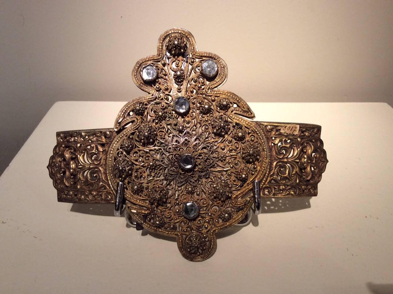 Belt buckle in gold-plated metal, filigree, glass cabochons.
This three-part belt buckle forming a floret is decorated on the sides with floral motifs, glass cabochons imitating the crystal adorn the central filigree part.
Balkans? 19th