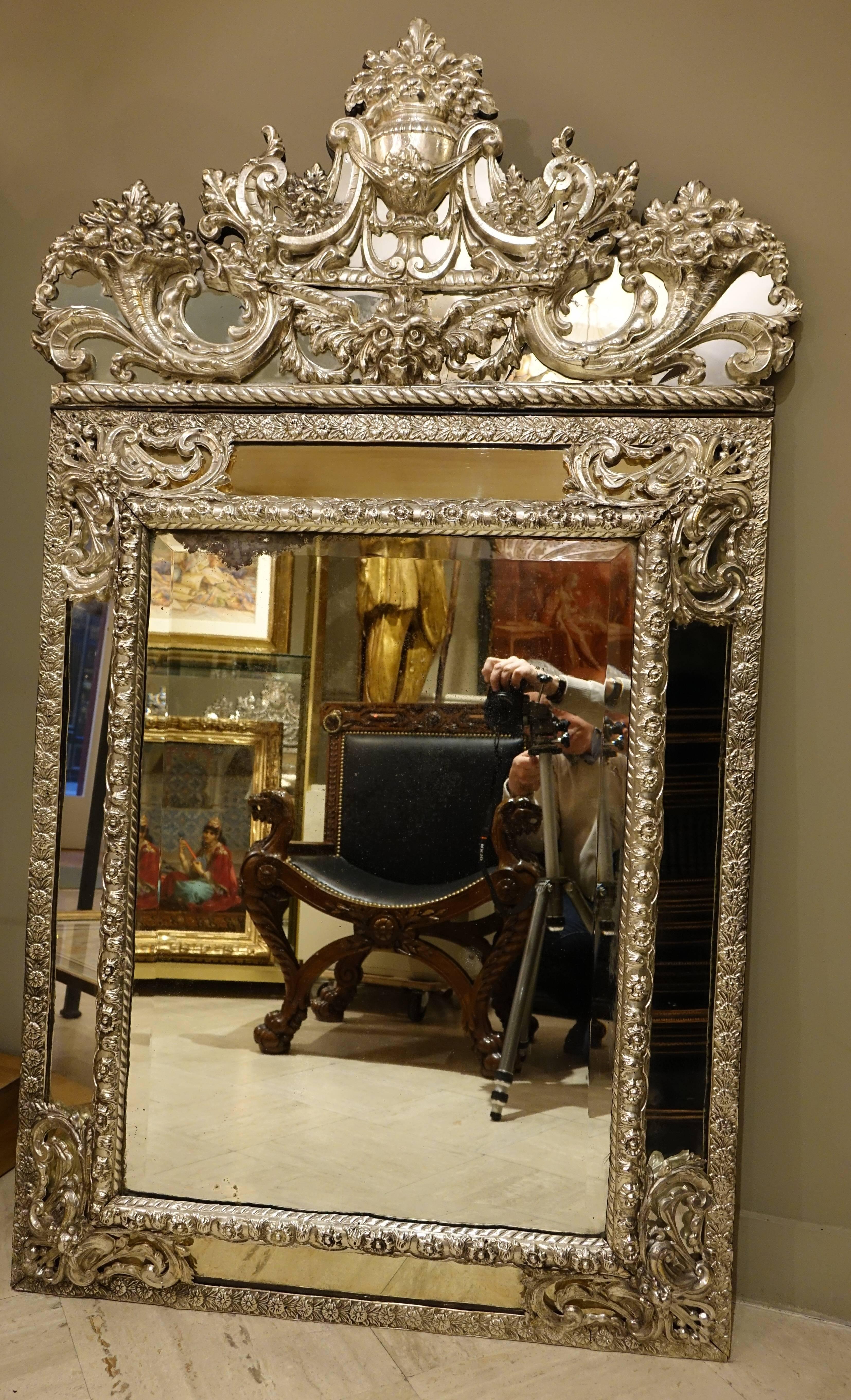 Mirror in Louis XIV style in embossed silver plated brass, France 19th century. 
The central mirror, the parclose mirrors on the sides, and the pediment have beveled edges. Decorated with scrolls, volutes, cassolettes and cornucopia 
Excellent