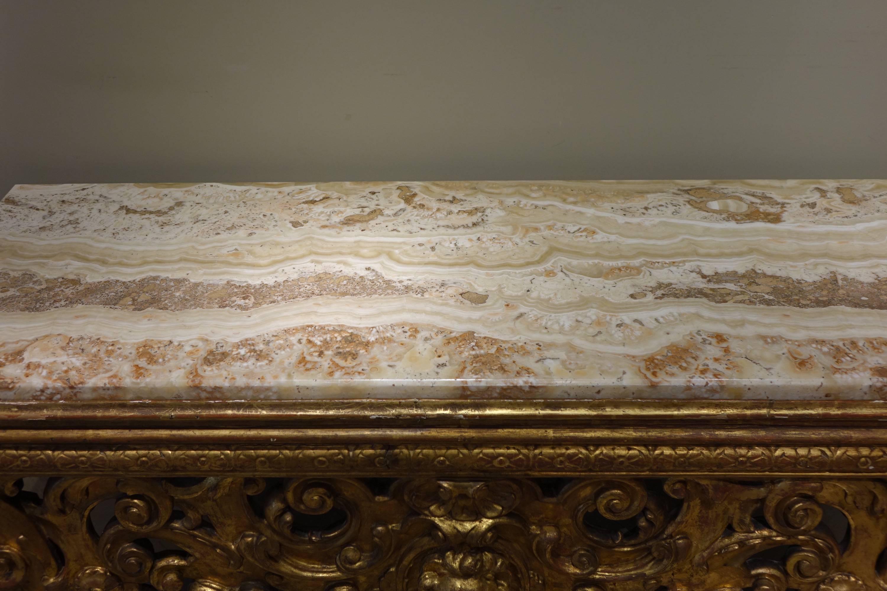 18th century Italian bench console formerly covered with fabric, replaced by an onyx slab, decorated with foliated scrolls, flower motifs and a lions head in its center.
The back is decorated with gilded wood with engravings. 
Probably Rome,