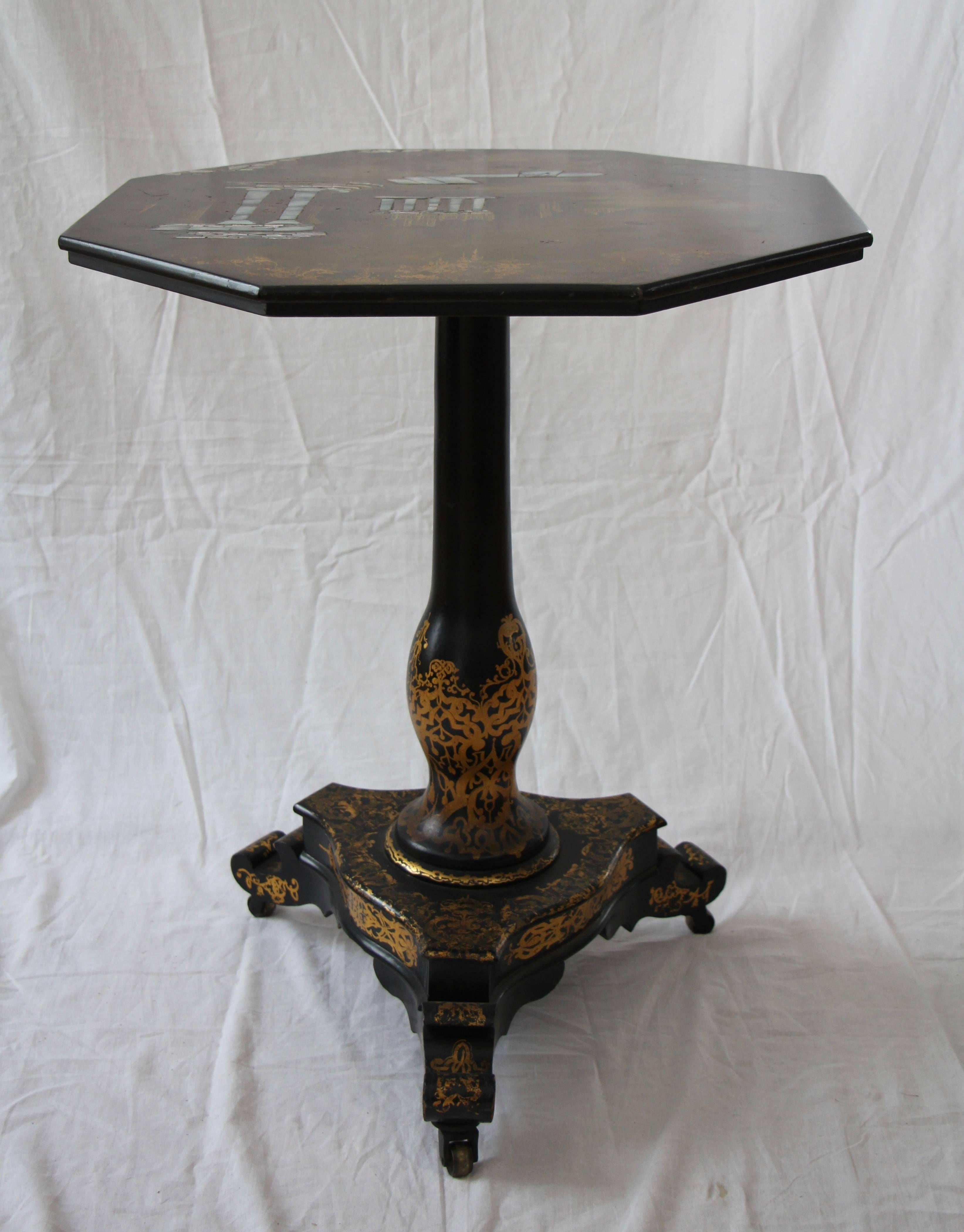 This English Victorian period tilt-top table has a octagonal top, decorated with polychrome paint and inlaid mother-of-pearl, depicting a landscape with antique temple architecture, all on a black and greenish lacquered ground, raised on a central