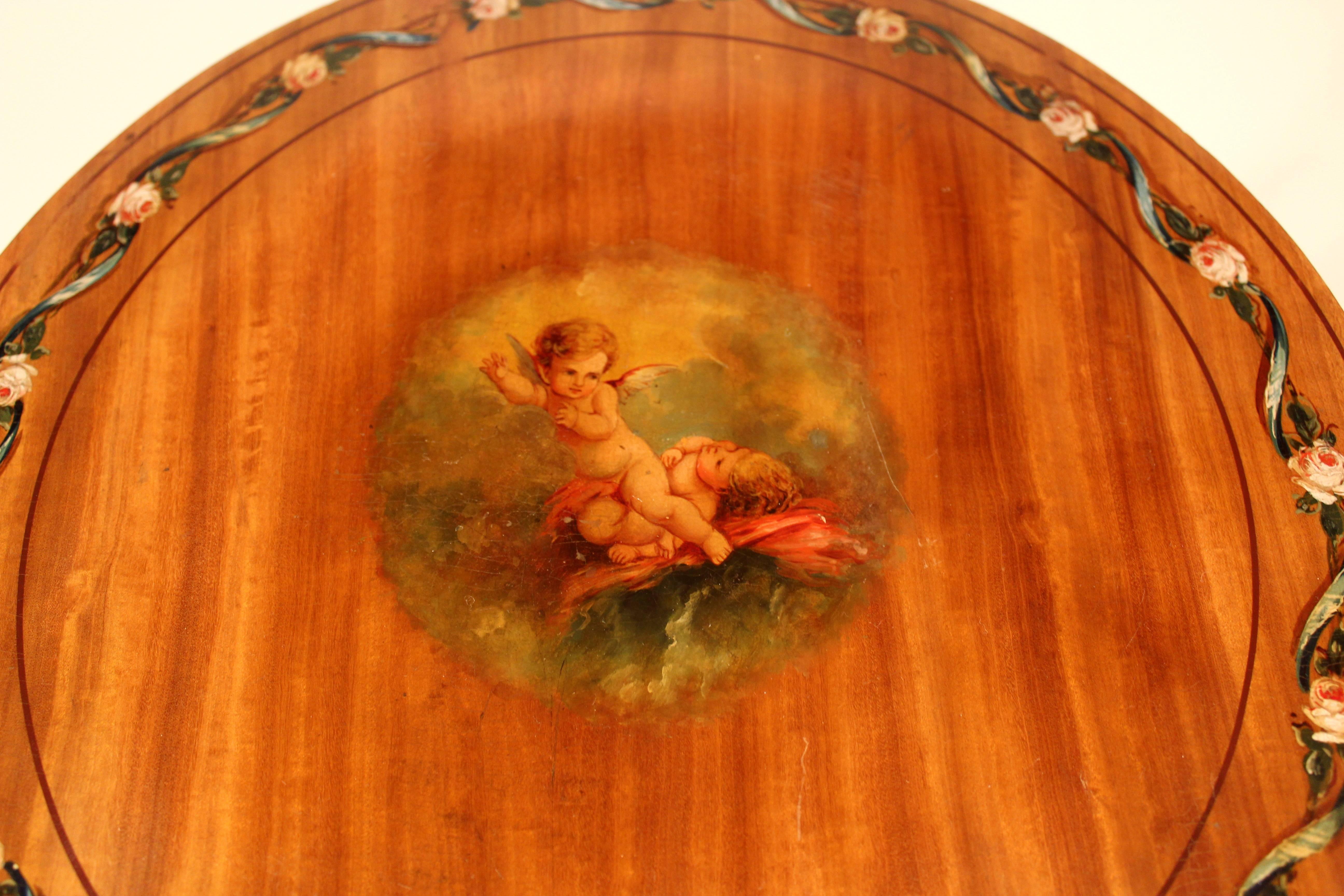 Satinwood working table with folding top. The round top painted with two putti in the center and a rose swag on the border. Raised on three tapering legs joined by a plate which shows  a presentation of  painted musical instruments. The apron and