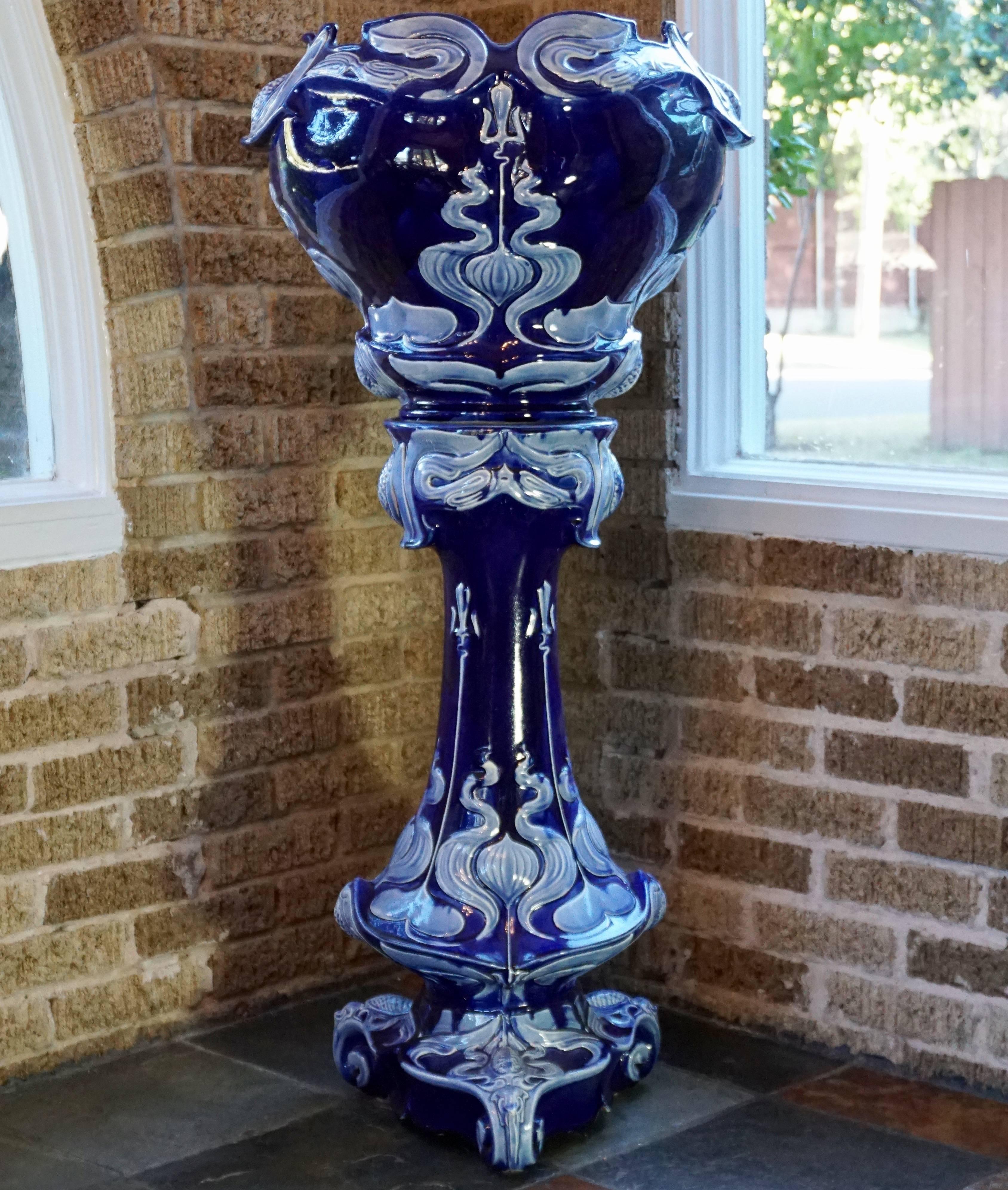 French Art Nouveau Vulcan cobalt pedestal jardinière, circa 1900. Decorated with a vivid array of colorful sculptured flowers and pine cones against a cobalt blue background with light scroll work.

Measures: Height 44 inches
Diameter 20.5