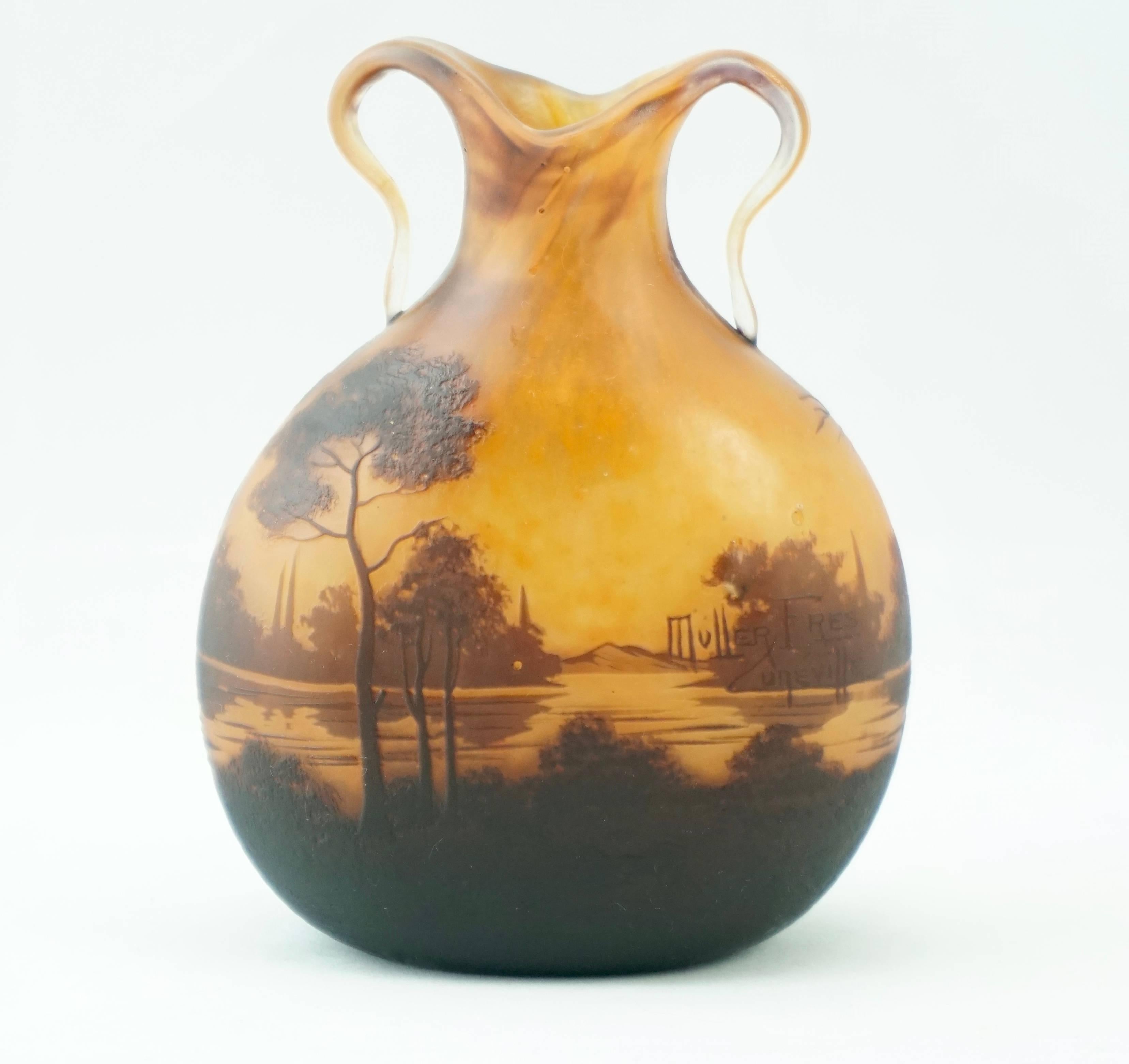 A sumptuous art nouveau French cameo lake landscape acid etched cameo vase with applied handles. Sunrise or sunset with a yellow orange background with engraved trees, bushes and mountains surrounding a pond or lake.

Signed in cameo: Muller Freres