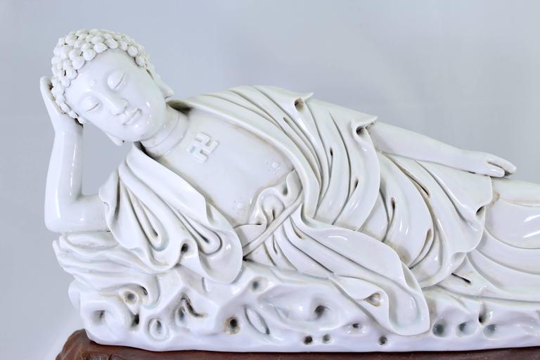 Shown a reclining or resting Siddhārtha Gautama, Shakyamuni Buddha with a wan emblem molded on the chest, wearing loosely draped robes falling in crisp folds, the face with a serene expression below spiky snail curls surrounding a smooth, rounded