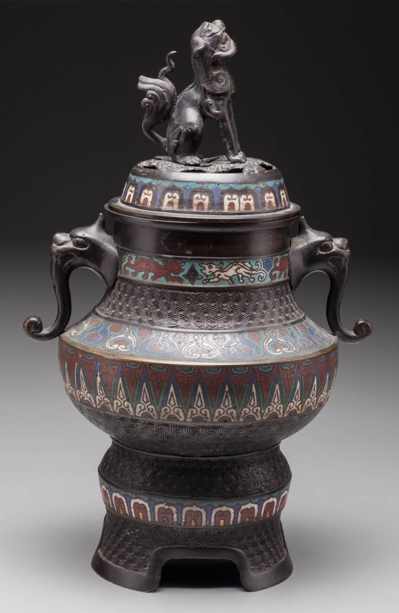 A Japanese Meiji period bronze and cloisonné censer, late 19th century.
Measures: 17 inches high (43.2 cm).

The censer having domed and reticulated lid with cloisonné banding and foo dog finial, figural mask handles, alternating bands of