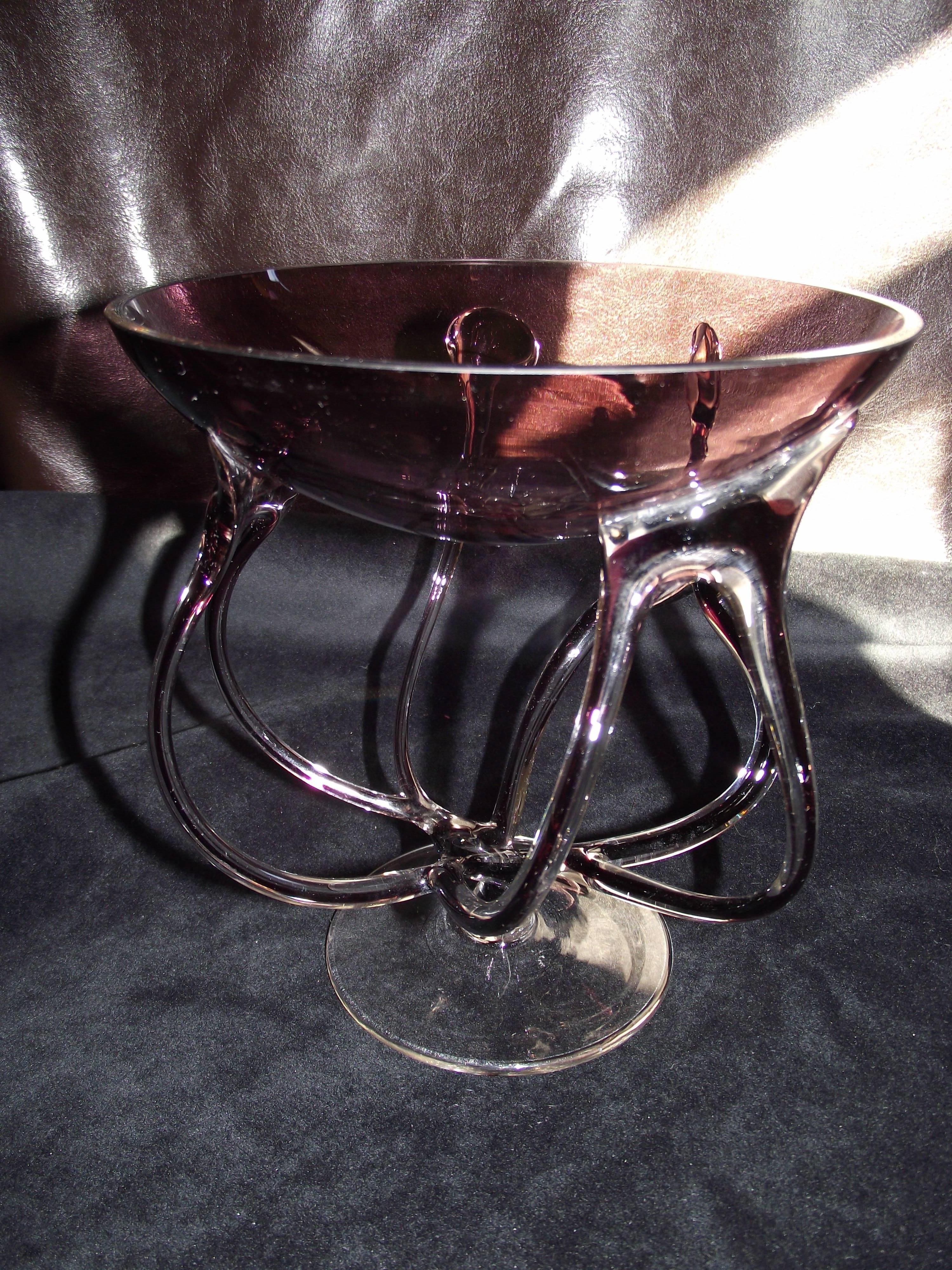 This one of a kind art glass piece has the free flowing lines of contemporary design with reference to a classic footed bowl. The bowl itself is a perfect shape of amethyst glass. The swirling legs area mix of clear and amber glass attaching to a