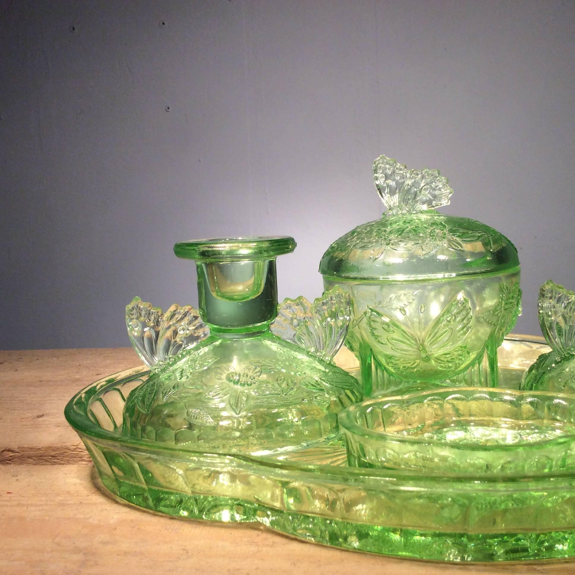Green glass dressing table set adorned with butterflies and floral motifs. The set comes with a tray, a pair of candlesticks, a large lidded pot and a ring dish.

Dimensions:

Ring dish: Length 9 cm, width 6.5 cm.

Candlesticks: Height 8 cm,