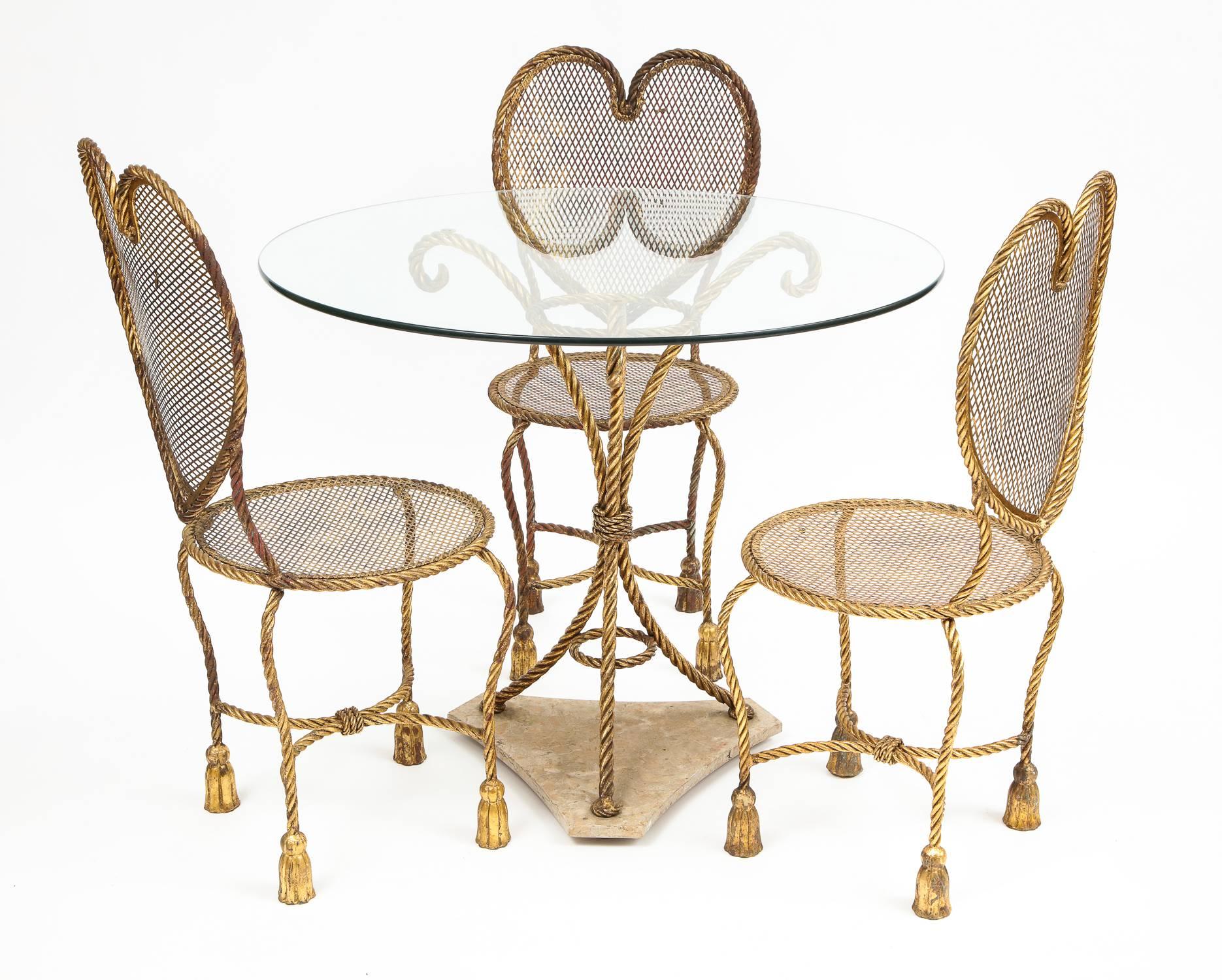 Italian gilt metal heart back Hollywood Regency tassel rope dining set 

Set includes three heart back woven gilt metal chairs with tassel rope motif and table with Italian travertine base and glass top.

Seat height: 17.5
