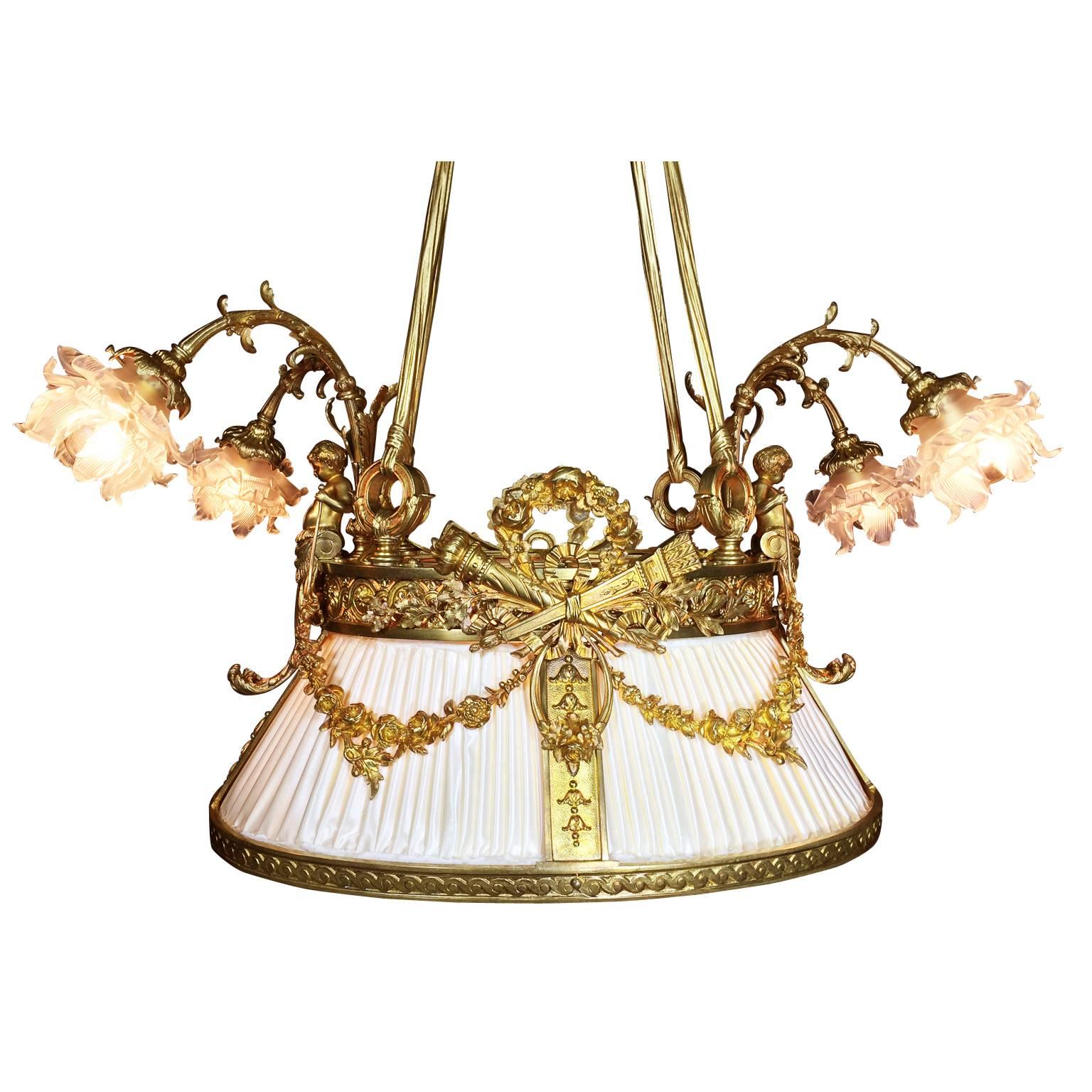 A fine and rare French 19th-20th century Belle Époque gilt bronze figural nine-light chandelier. The ovoid shaped frame, in the form of a hanging shade, surmounted with a gilt bronze Putto at each end, both holding floral wreaths below a two-light