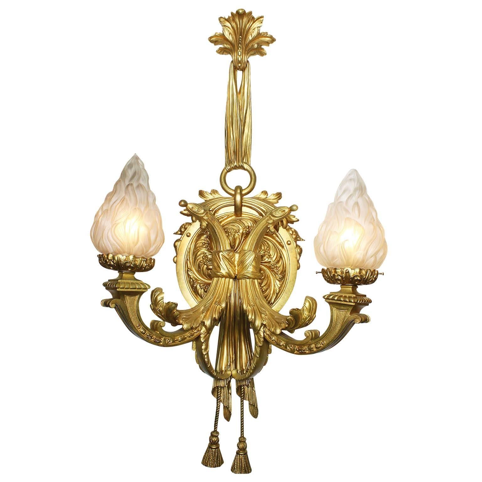 A fine and impressive pair of French 19th-20th century Louis XVI style gilt bronze two-light wall sconces, each lumière with a circular foliate backplate suspended by a ribbon tied ornament issuing two-arm form cornucopias holding frosted glass