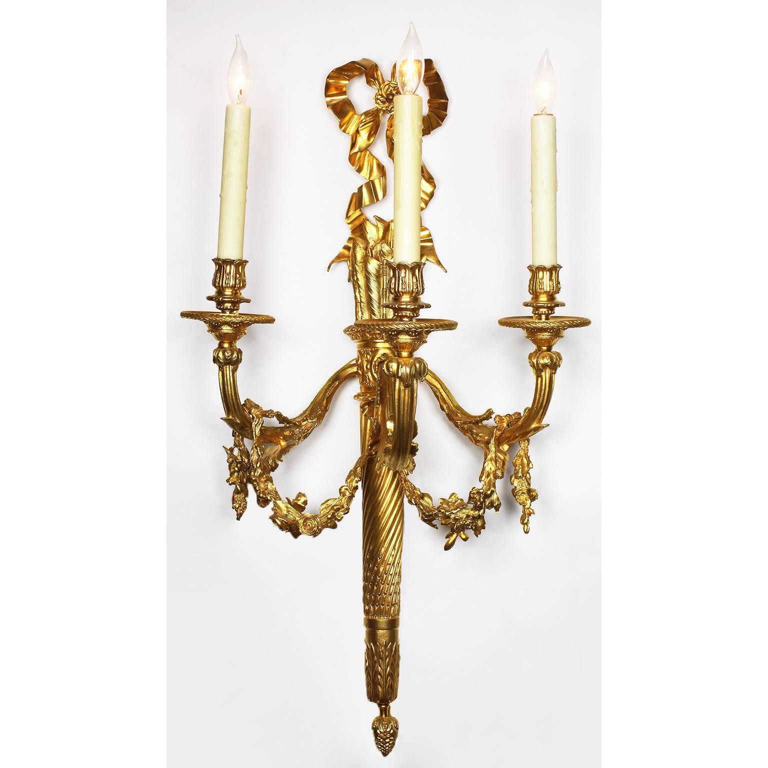 A fine pair of French 19th-20th century Louis XVI Style Gilt-Bronze Three-Light Wall Sconces. The back plate in the form of a torch with three scrolled candle arms protruding from the center frame, each arm conjoined floral wreath swags. The upper