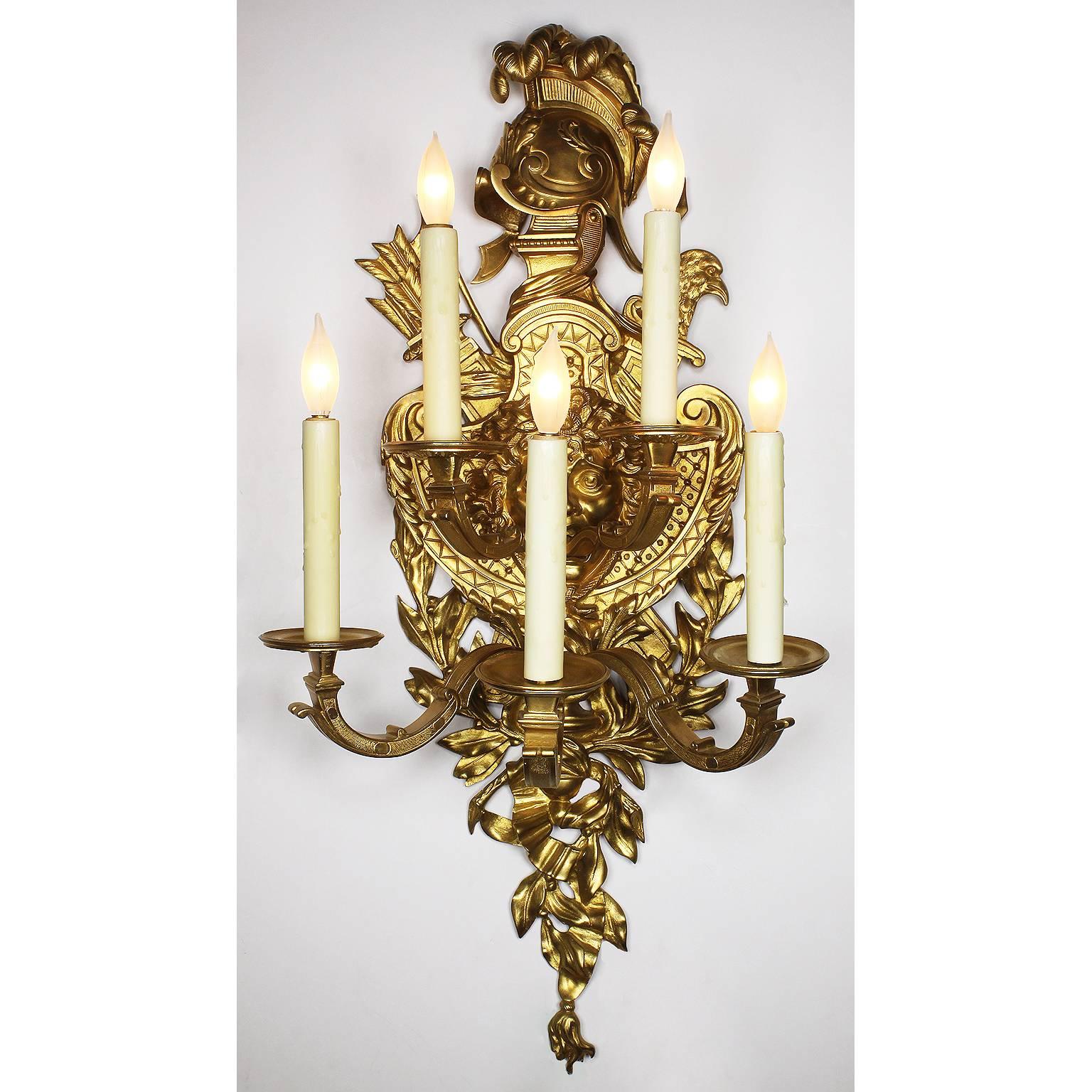 A pair of French 19th-20th century Regence style gilt bronze five-light figural wall lights, each with a ribbon-tied back-plate suspending a plumed helmet and centred by a Medusa mask shield with military trophies issuing two acanthus cast