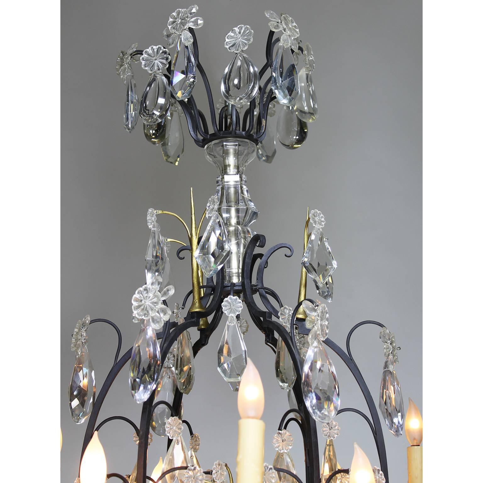 A French 19th-20th century Louis XV style wrought iron and parcel-gilt eighteen-light crystal (cut-glass) chandelier. The black painted frame with twelve candle arms and six interior parcel-gilt cups, all electrified, surmounted with crystal