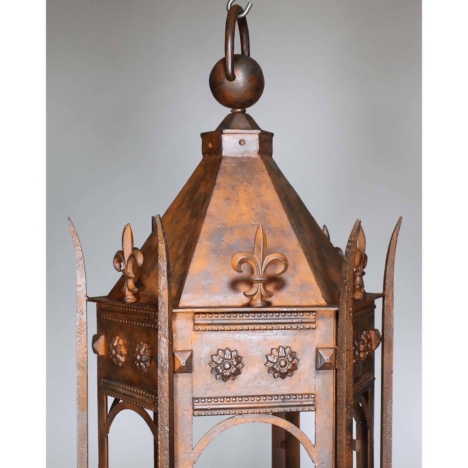 A set of four hexagonal French Louis XV style wrought iron hanging lanterns, after the antique. The elongated body surmounted with fleur-de-lis', floral rosettes and scrolled decorative elements, in a rusted brick finish, circa late 20th century.