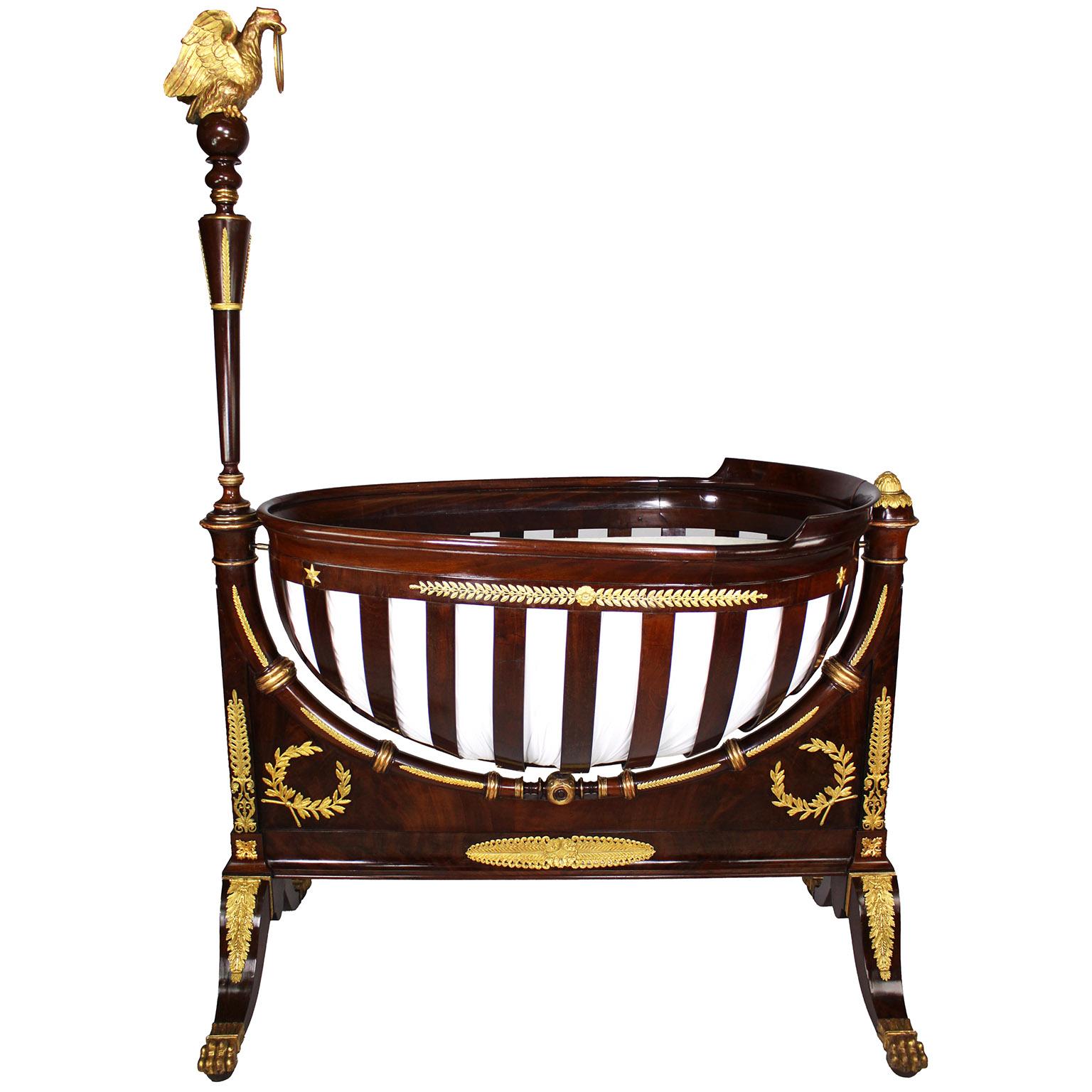 A Very Fine French or Russian 19th Century Empire Style Mahogany, Ormolu Mounted and Parcel-Gildwood Carved Crib & Bedding. The egg-shaped oblong rocking cradle supported at each end by columnar standards.The cradle base conforms to the rocking