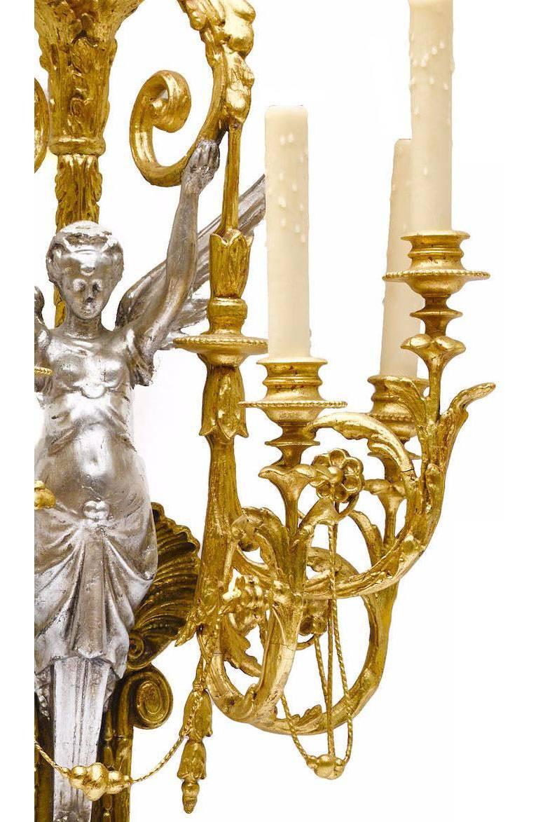 A fine and rare pair of French neoclassical style 19th century figural parcel gilt and silver carved wood and Gesso six-light wall sconces, each centered with giltwood carved seashell behind a silver-leaf carved wood figure of a Victorious Fairy