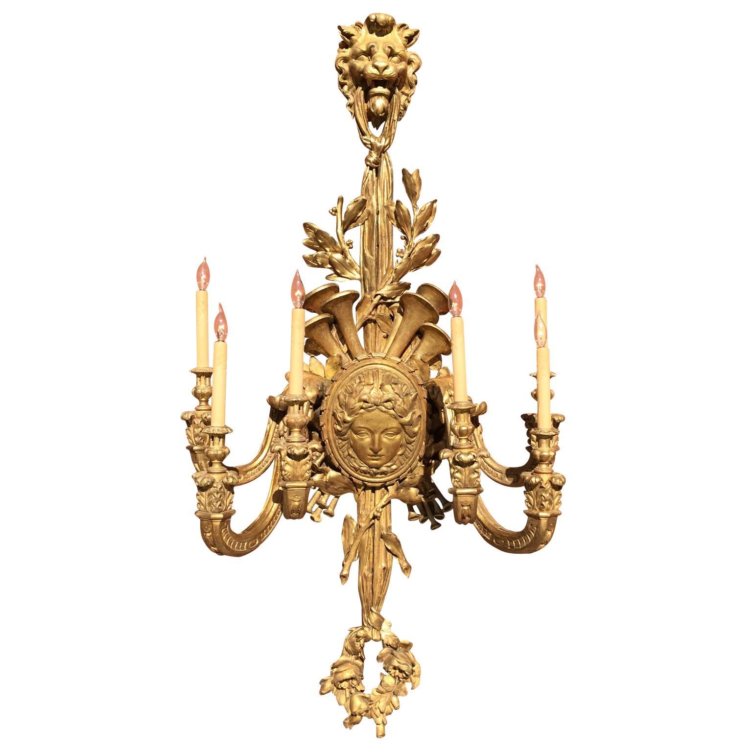 Importante y palaciega pareja de apliques de seis luces tallados en madera dorada del siglo XIX, estilo Luis XV francés, con seis brazos de vela en volutas que emanan de un medallón central con una máscara femenina y rematados con instrumentos