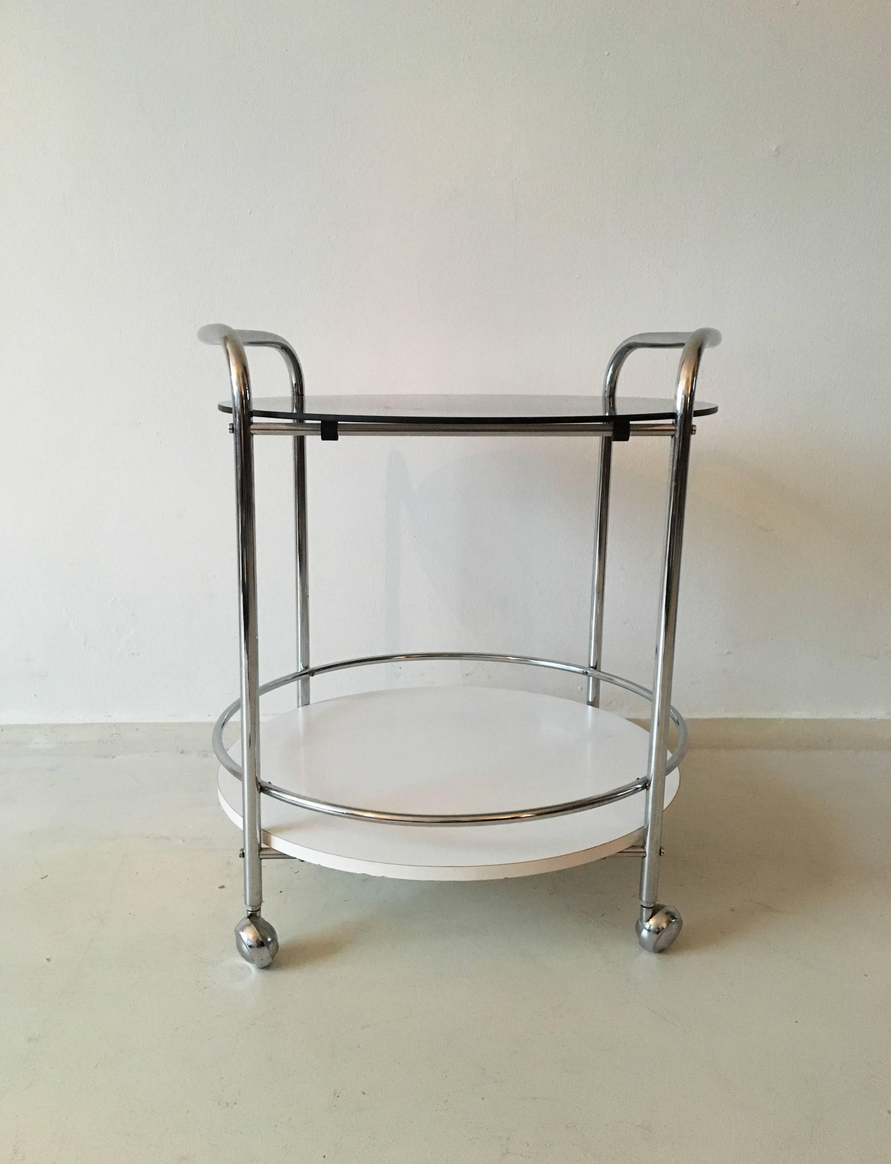 This Mid-Century Modernist trolley or bar cart consists of a chrome-plated tubular frame with one smoked glass top and one white veneered/laminated top in wood. Overall in a very good condition with some wear to the glass.
