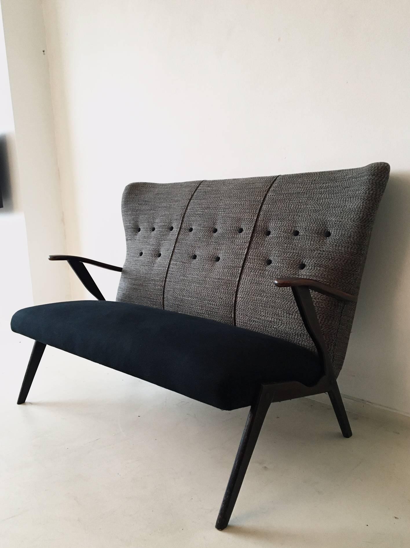 This small grey fabric sofa was designed during the 1950s in the style of Ercol. It was manufactured in Belgium. The sofa features a wooden frame, and a recently re-upholstered dark black fabric seat and dark grey backrest. Although this sofa is in