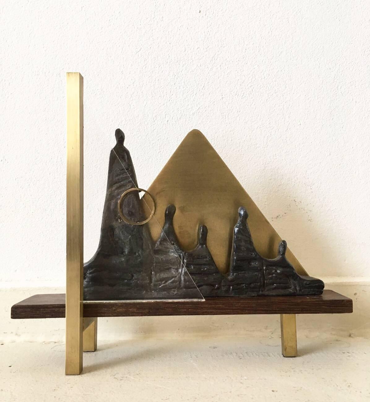 This stunning and unique piece of work was created by Judith Braun in the late 1980s. 

It features people in bronze that sit on a wengé wooden base. Behind them, a triangular form in brass probably simulating a pyramid.
At the front, a brass