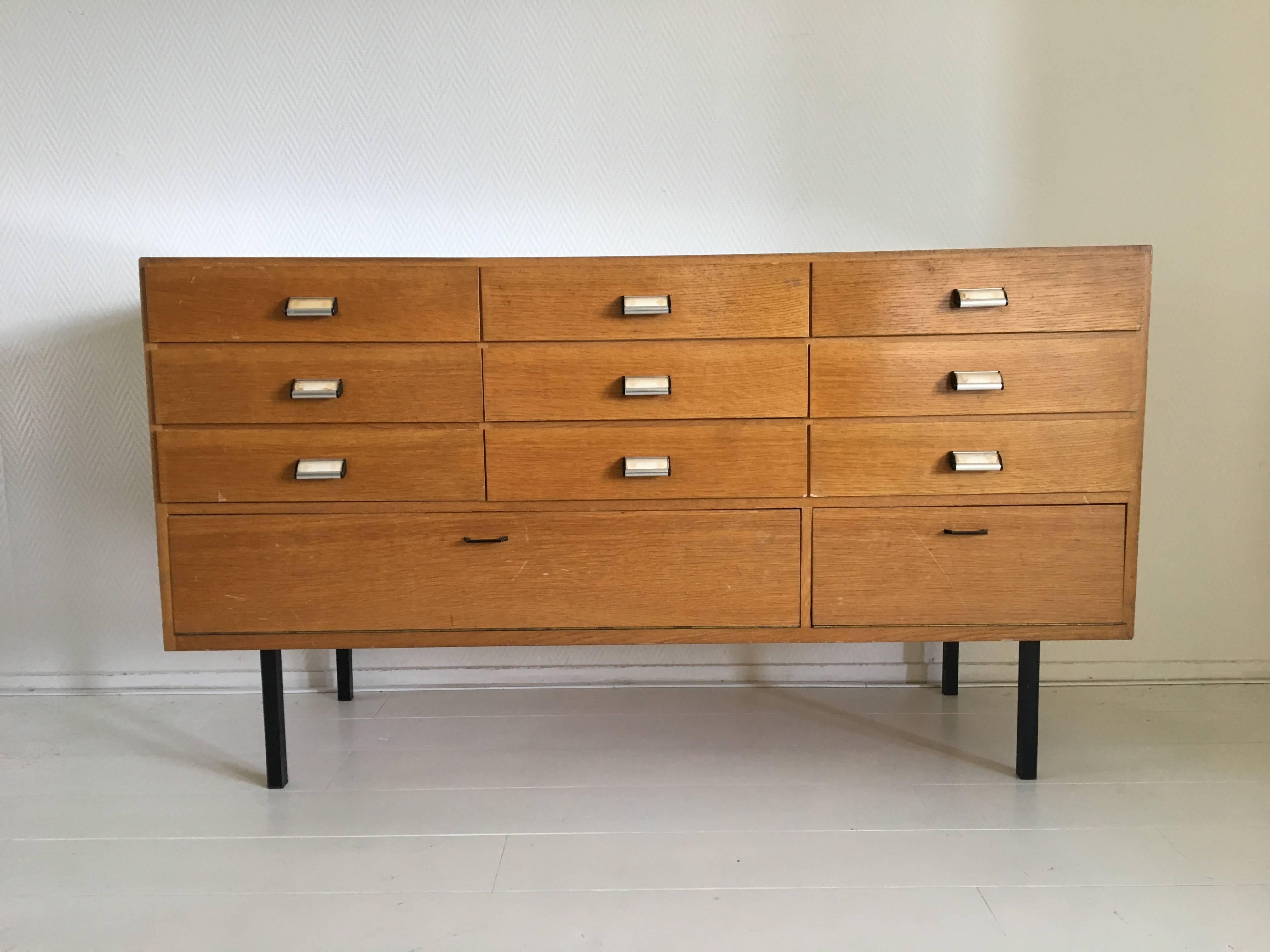 This Industrial looking piece was designed and manufactured in Europe, circa 1950s. It features an oak and thick glass body with black metal feet. It's nine drawers feature very nicely looking handles. Underneath the drawers there are two drop down