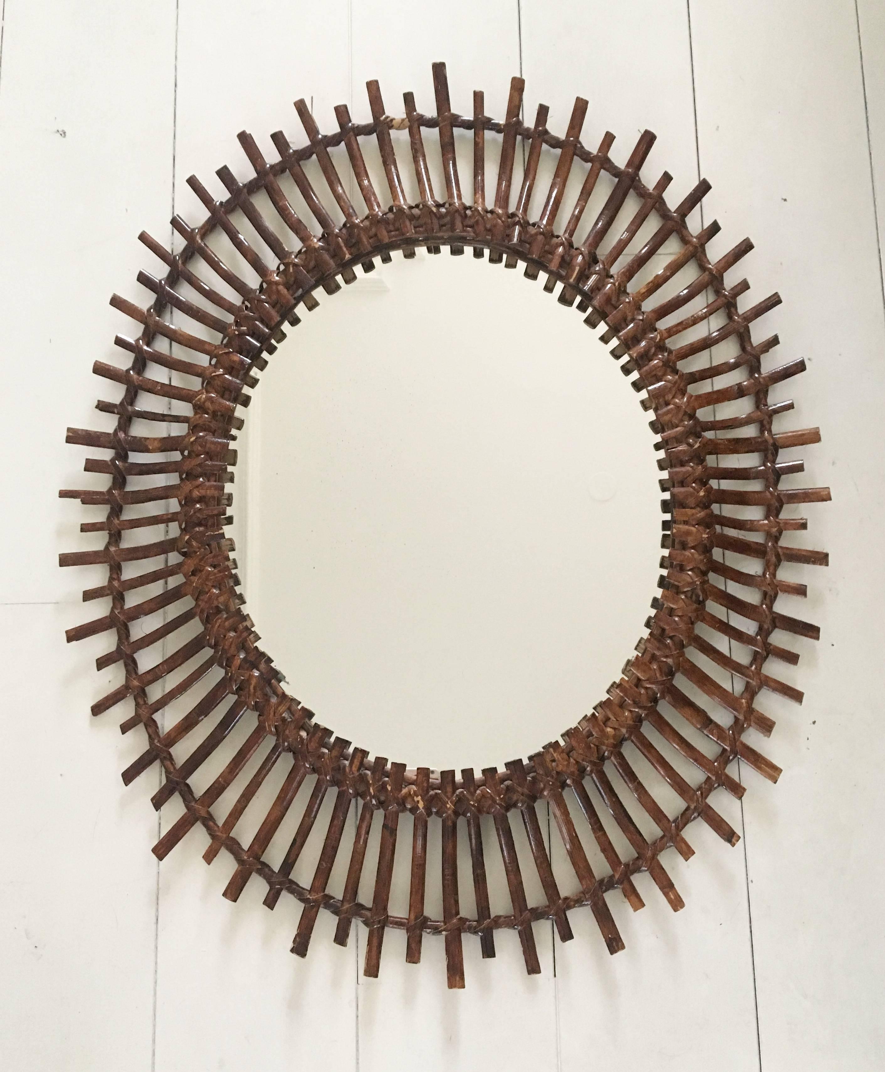 Elegantly handcrafted rattan or wicker sunburst mirror with oval shape, in the style of Franco Albini. The mirror features a row of curved beams in different lengths and is finished with a coat lacquer. The back of the mirror, features a leather