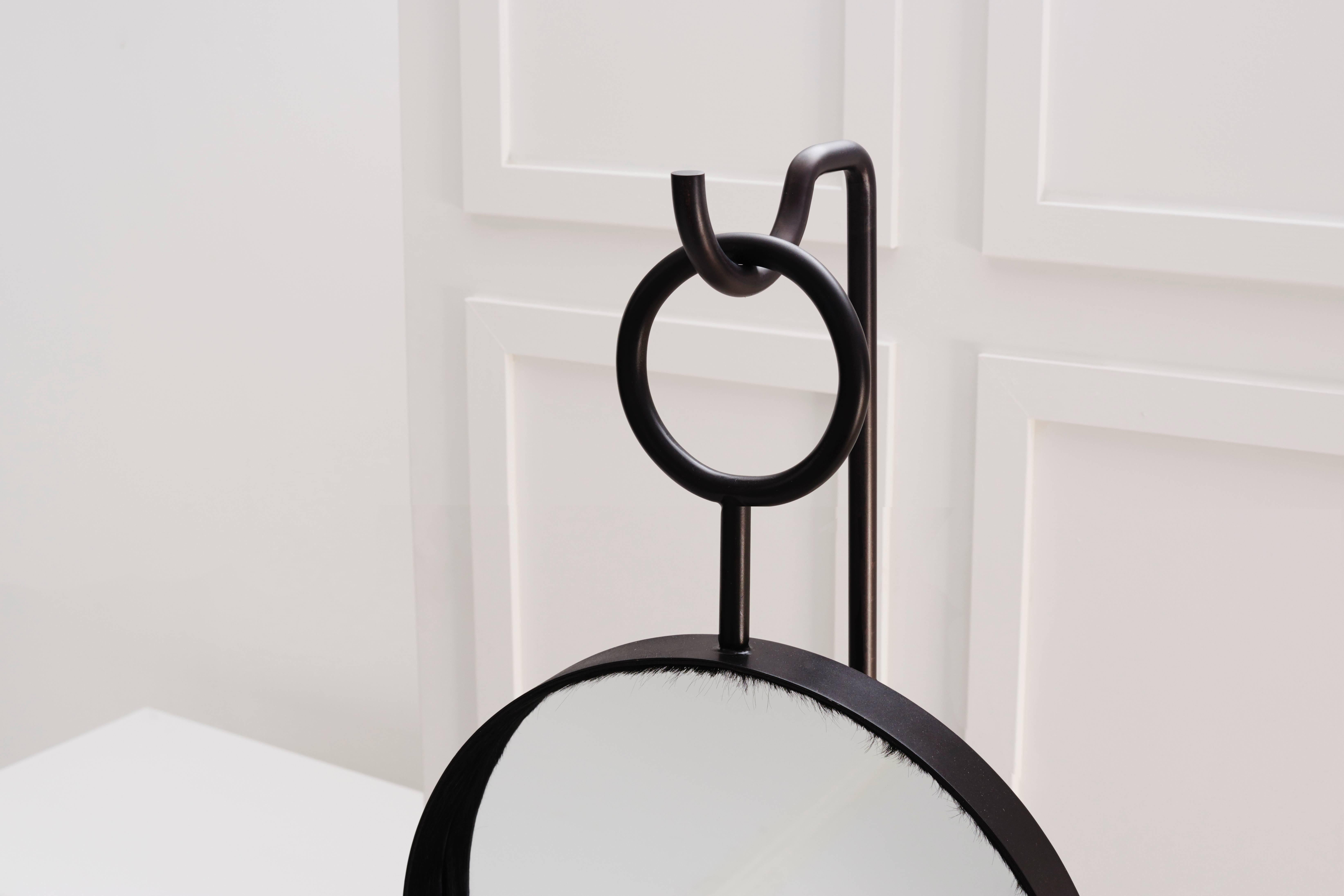 A round mirror, inset deeply in its frame, hangs on a loop from the hook of its accompanying base. This sculptural piece is hand finished in either a rich warm black or antique brass.