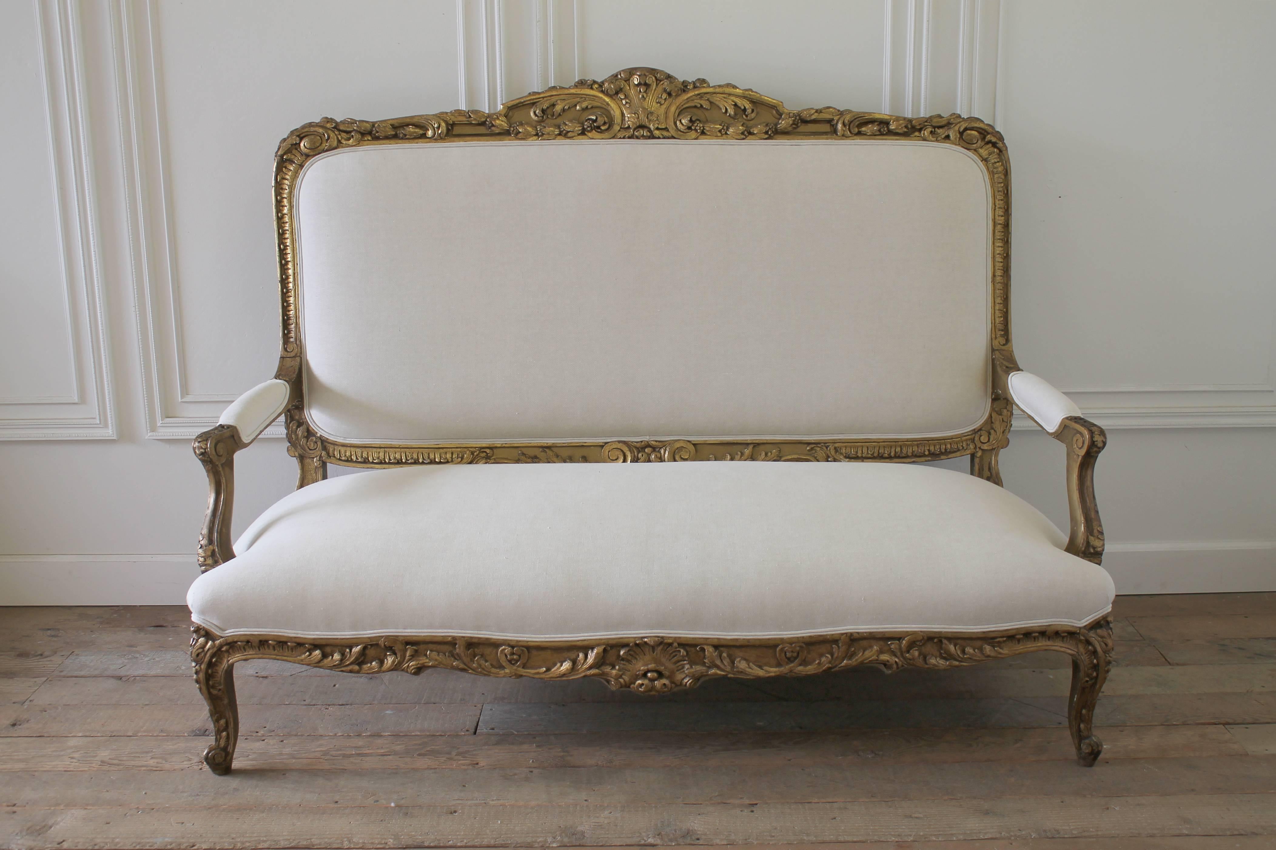 Gorgeous giltwood settee has intricate carved details of scrolls and leaf and berry motifs. We have reupholstered this in our 100% pure Belgian linen, color is oatmeal, or off-white. Finished with a double welt, this settee has original spring seat,