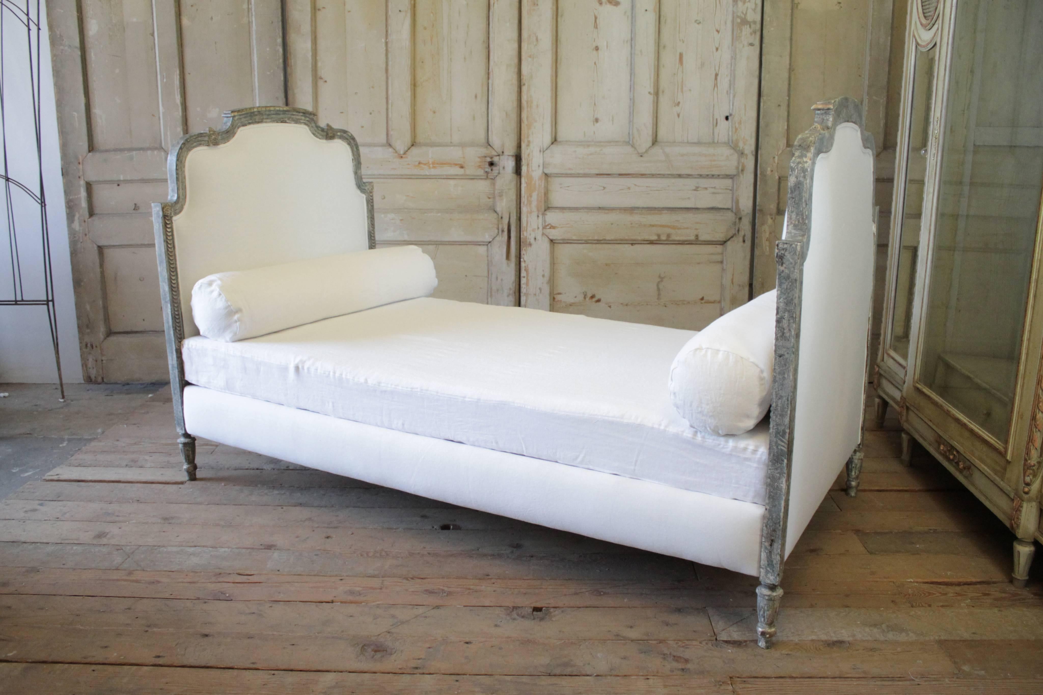 Beautiful original painted Louis XVI style daybed in a grey blue weathered patina, with hints of a silver leaf painted accents. We reupholstered this in a thick white Belgian linen and finished with a double welt edge. The rails for the bed were