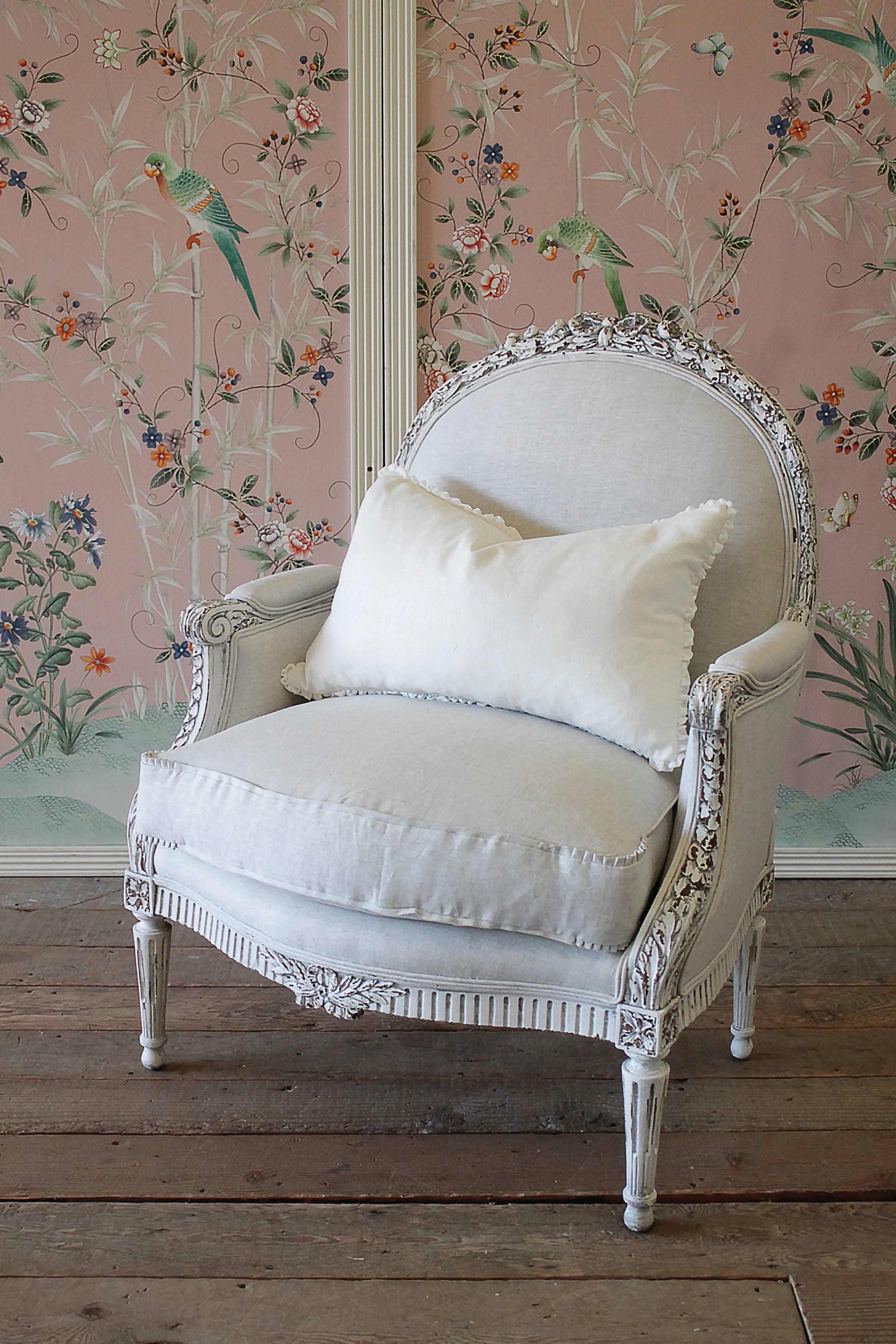 Early 20th century carved and painted Louis XVI style bergere chair painted in our soft oyster white finish, distressed edges and finished with an antique glazed patina. The painted finish is an off-white, with subtle hues of a grey tone. Large