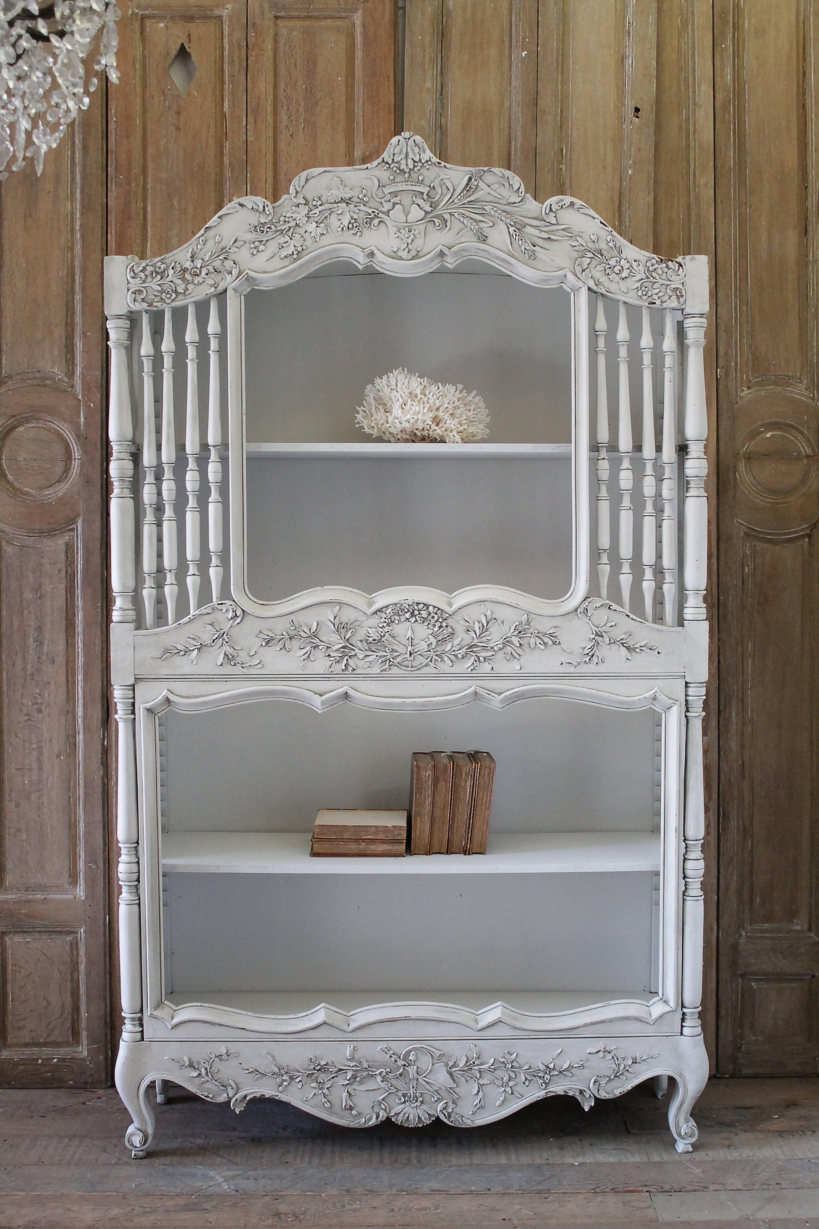 19th century painted Louis XV style display bookcase with beautiful carvings of a shield crest and crown with vines and leaves trailing across the top. The center carvings are of laurel leaves trailing away from a floral crown. Three adjustable