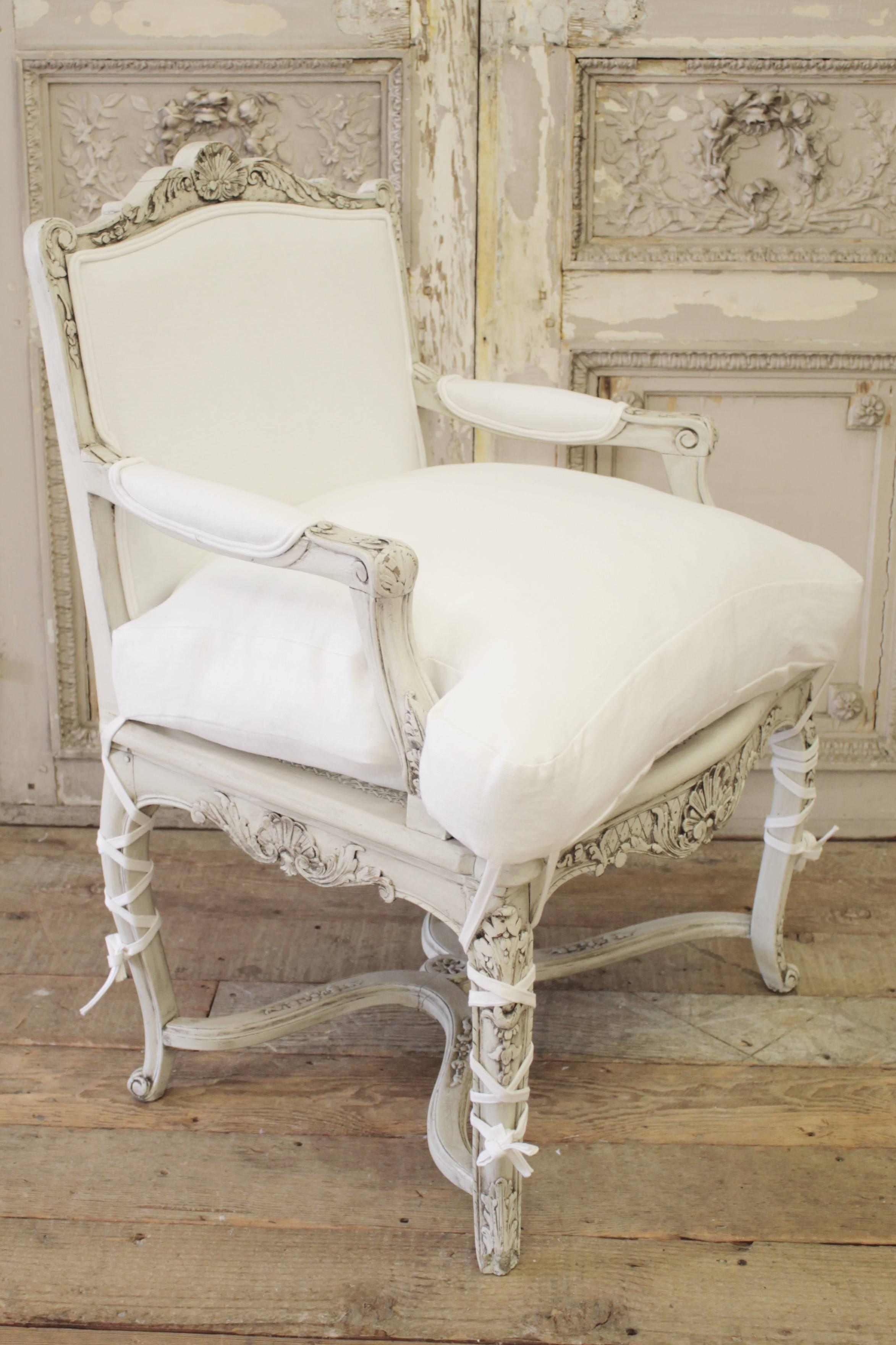 Painted in a soft oyster with subtle distressing, this hand-carved chair has the original cane seat, upholstered back and arms in 100% pure Belgian linen. The seat is slip covered down, with a ballet tie leg. The cushion cover has a zipper and can
