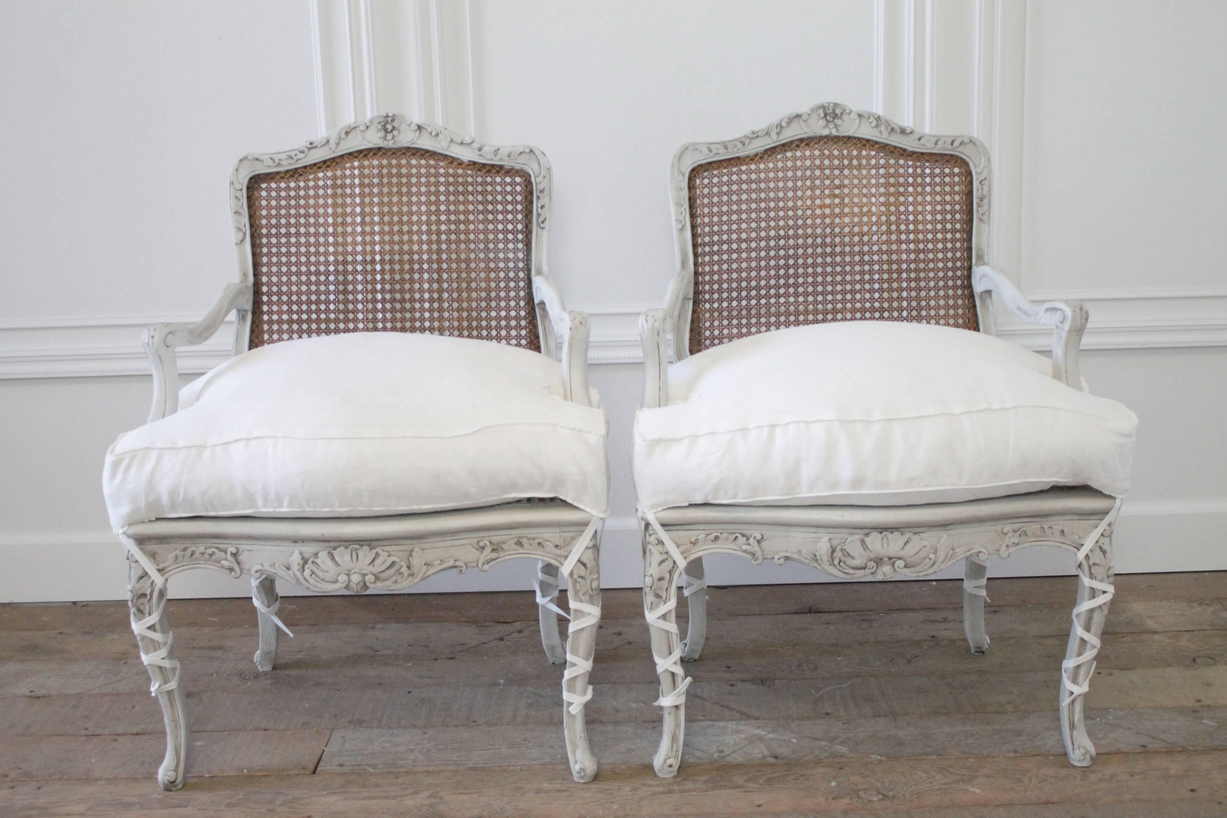 Painted in our oyster white, with the original natural cane, and an antique glazed patina. These carved armchairs have a carved flower with trailing scrolling leaf designs. The cane was left original, and the one of the chairs has been repaired, the
