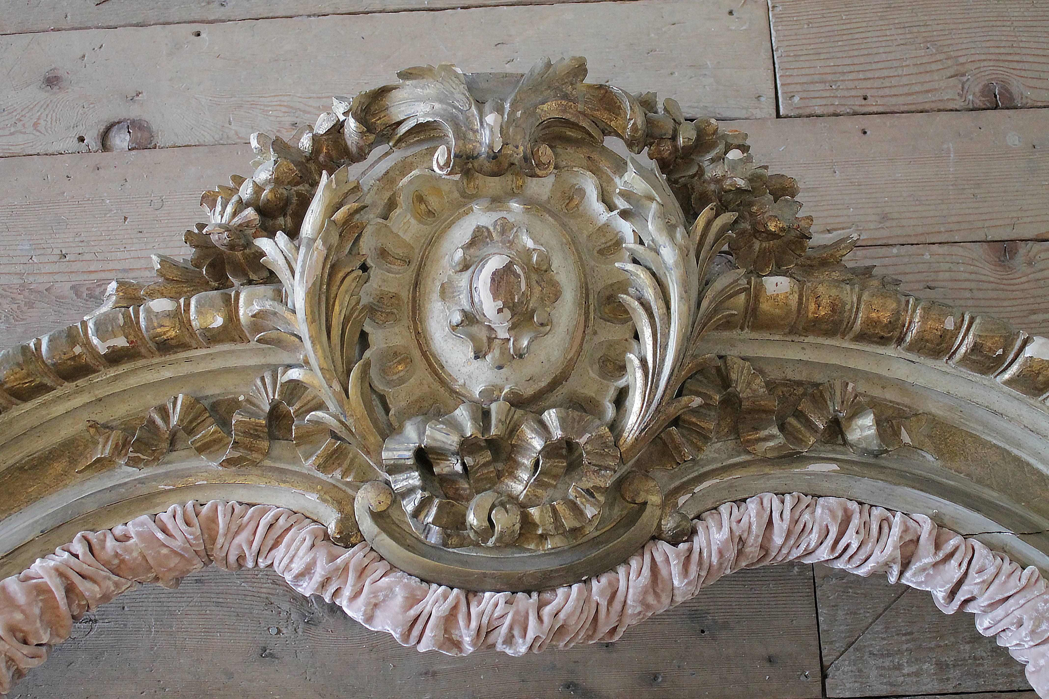 Beautiful painted and gilded French bed crown with large carved medallion crest and ribbon carvings. The top has trailing flowers and roses swaging down each side. The bottom has a vintage pink crushed velvet rusching. There are mounting brackets