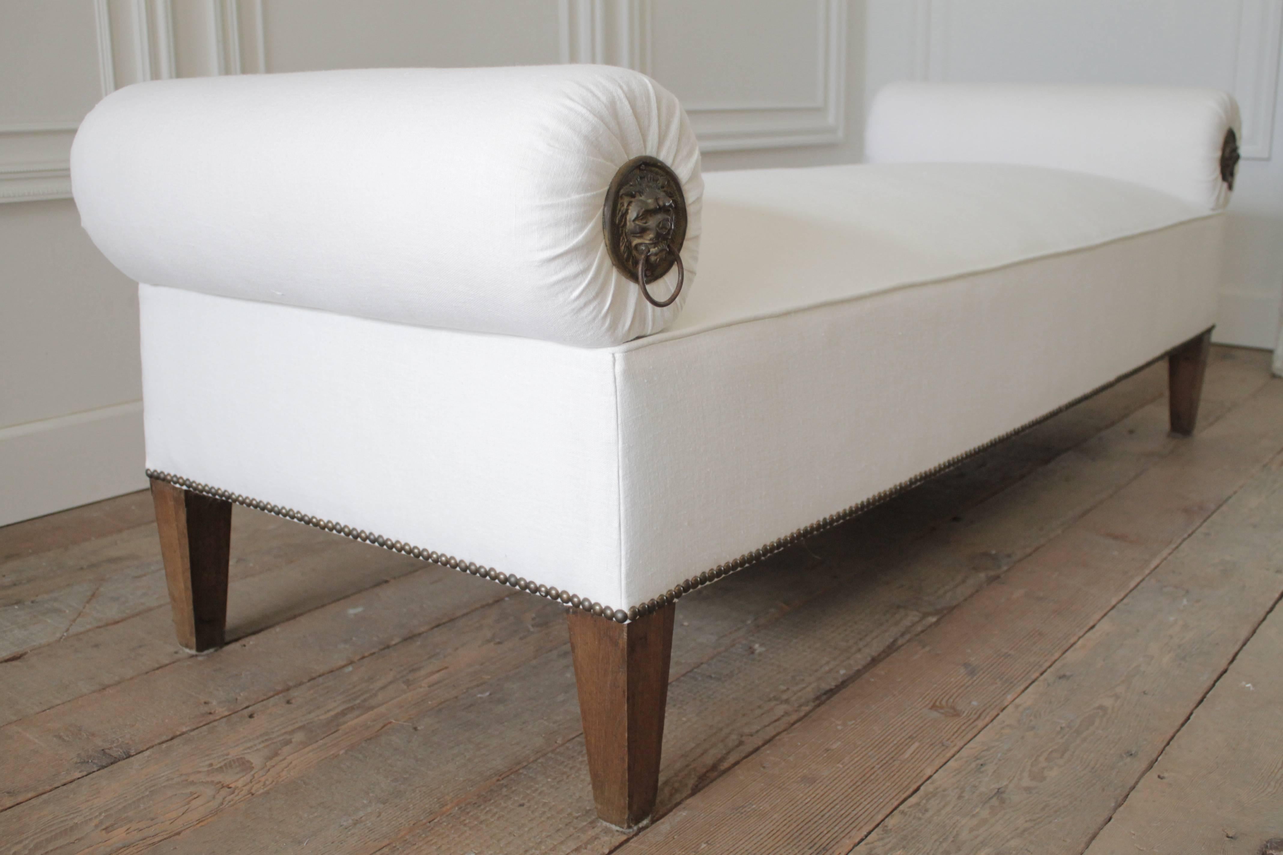 Beautiful custom designed bench with solid oak legs and antique brass nailhead trim. We reupholstered this in a soft white heavy weight Irish linen. The round bolsters feature lions head knocker hardware on one side. These bolsters are removable for