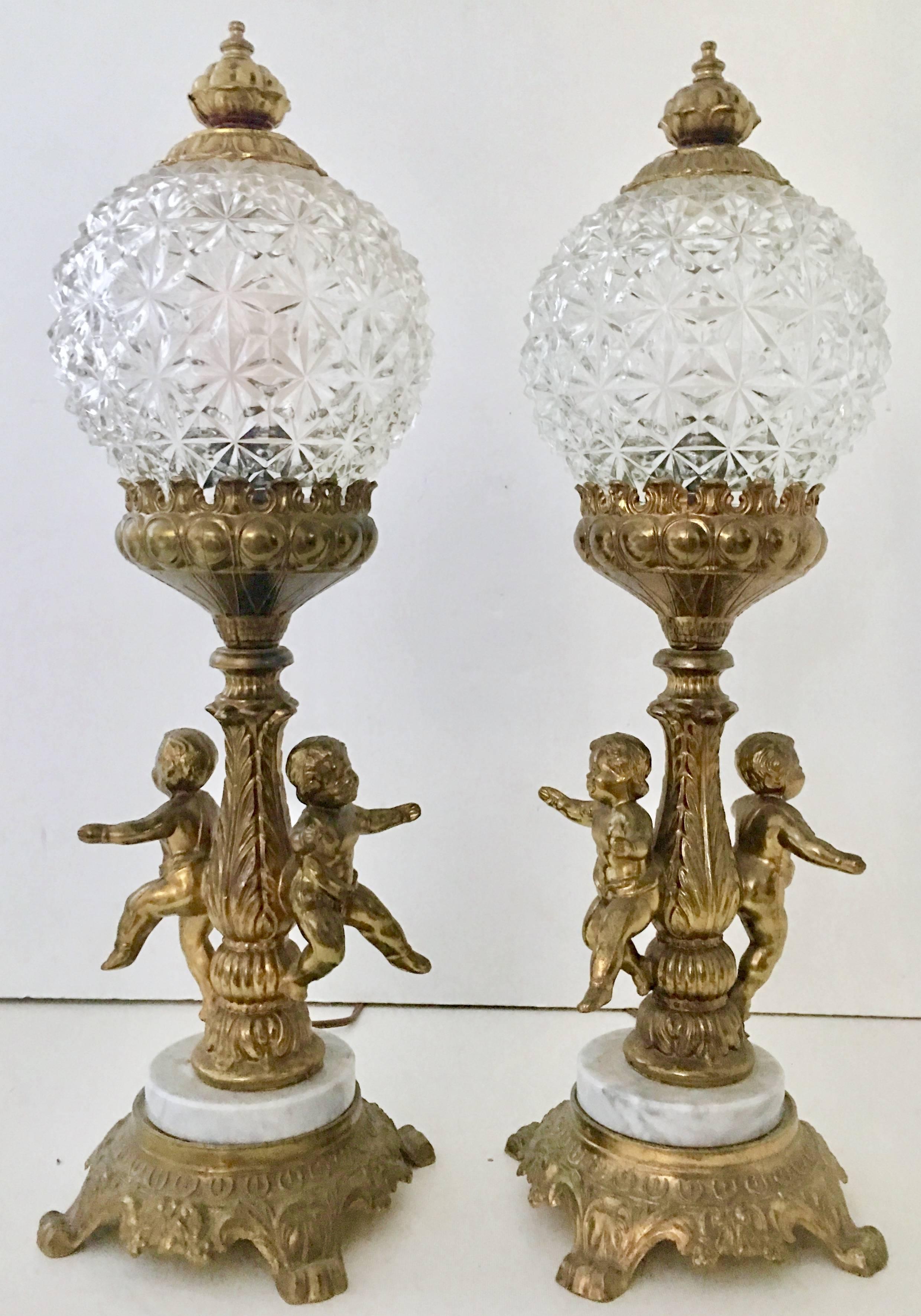 Antique pair of French style bronze ormolu and marble mounted putti electrified oil lamps. These fantastic double putti or cherub converted oil lamps feature a cut glass diamond pattern round globe with brass finial shade. Both cut-glass shades are