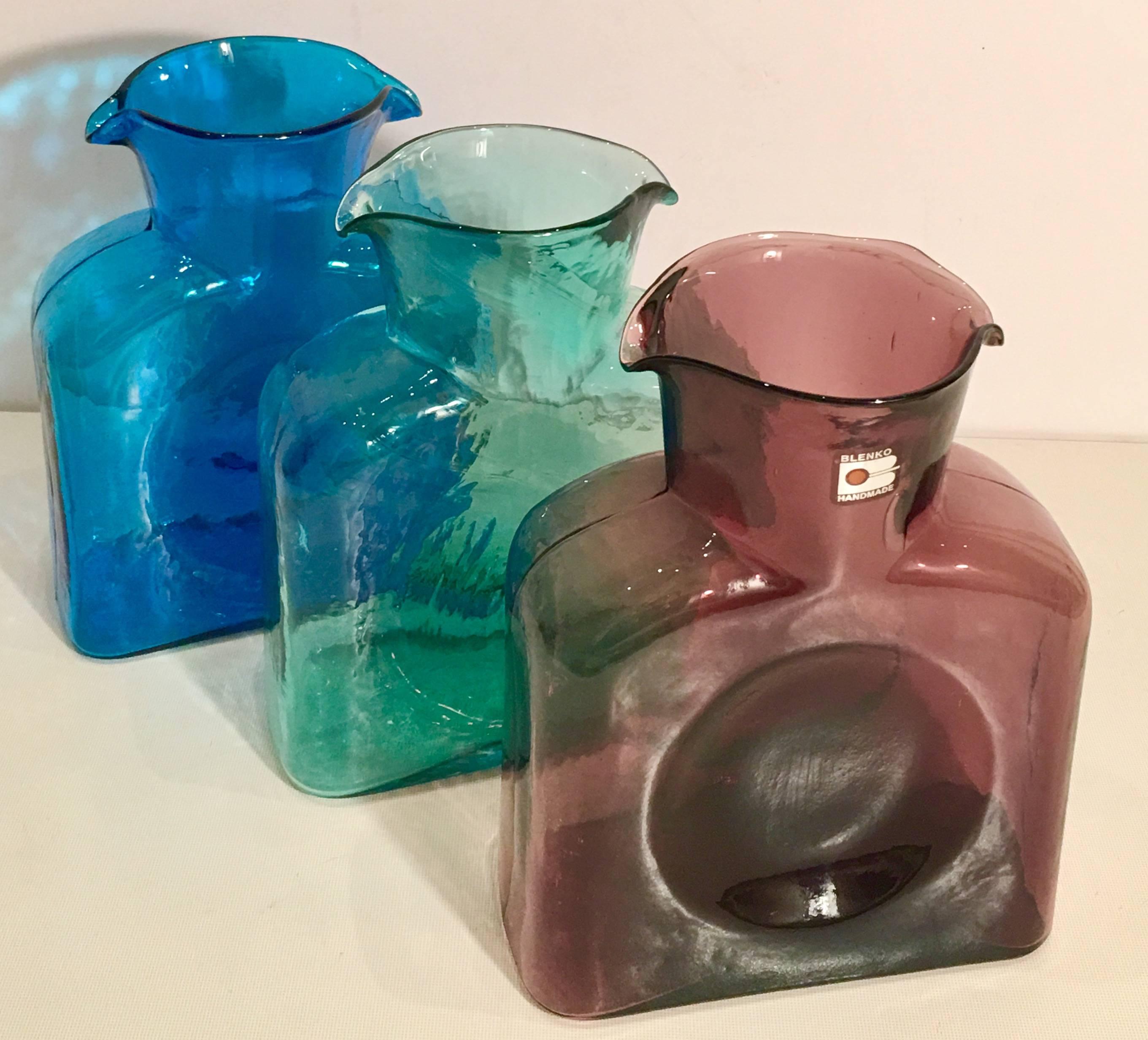 Mid-Century Blenko glass trio of double spouted pitchers-jugs. This trio features, one blue, green and amethyst. The amethyst pitcher has the original Blenko glass manufacturer sticker intact.
