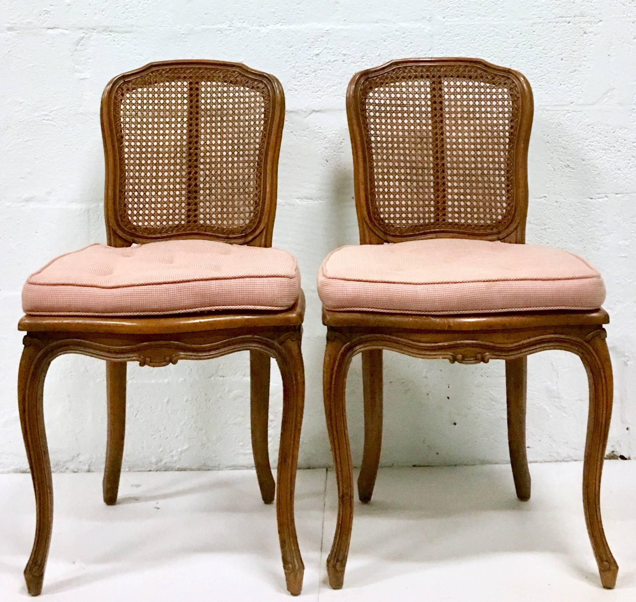 Pair of French Provincial style double cane wicker and carved wood chairs. Features double cane seat and back. Each chair includes a pink gingham check cotton blend and button tufted slip cover seat cushion with Velcro ties.. Seat height, 16