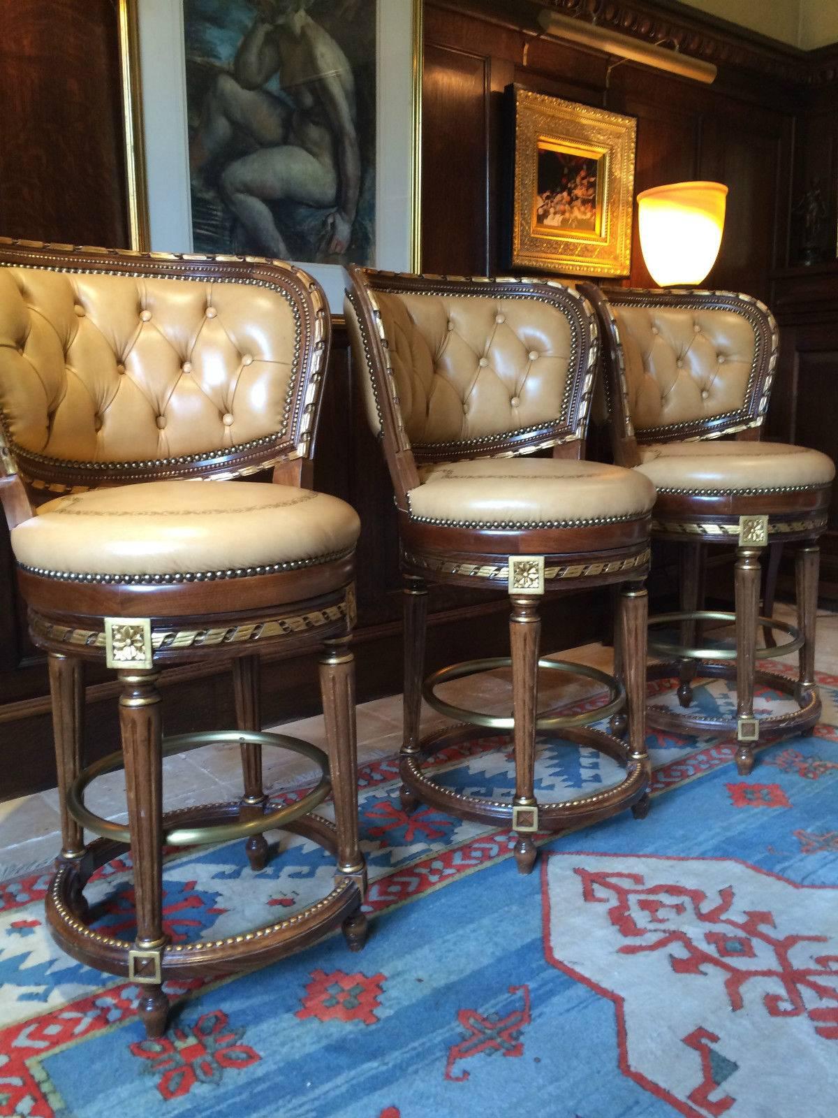 A trio of French Louis XV style rotating high back bar stools, with parcel-gilt mahogany frames and tan leather button back upholstered back supports, tan with gilt leather overstuffed upholstered rotating seats, fluted column legs united by