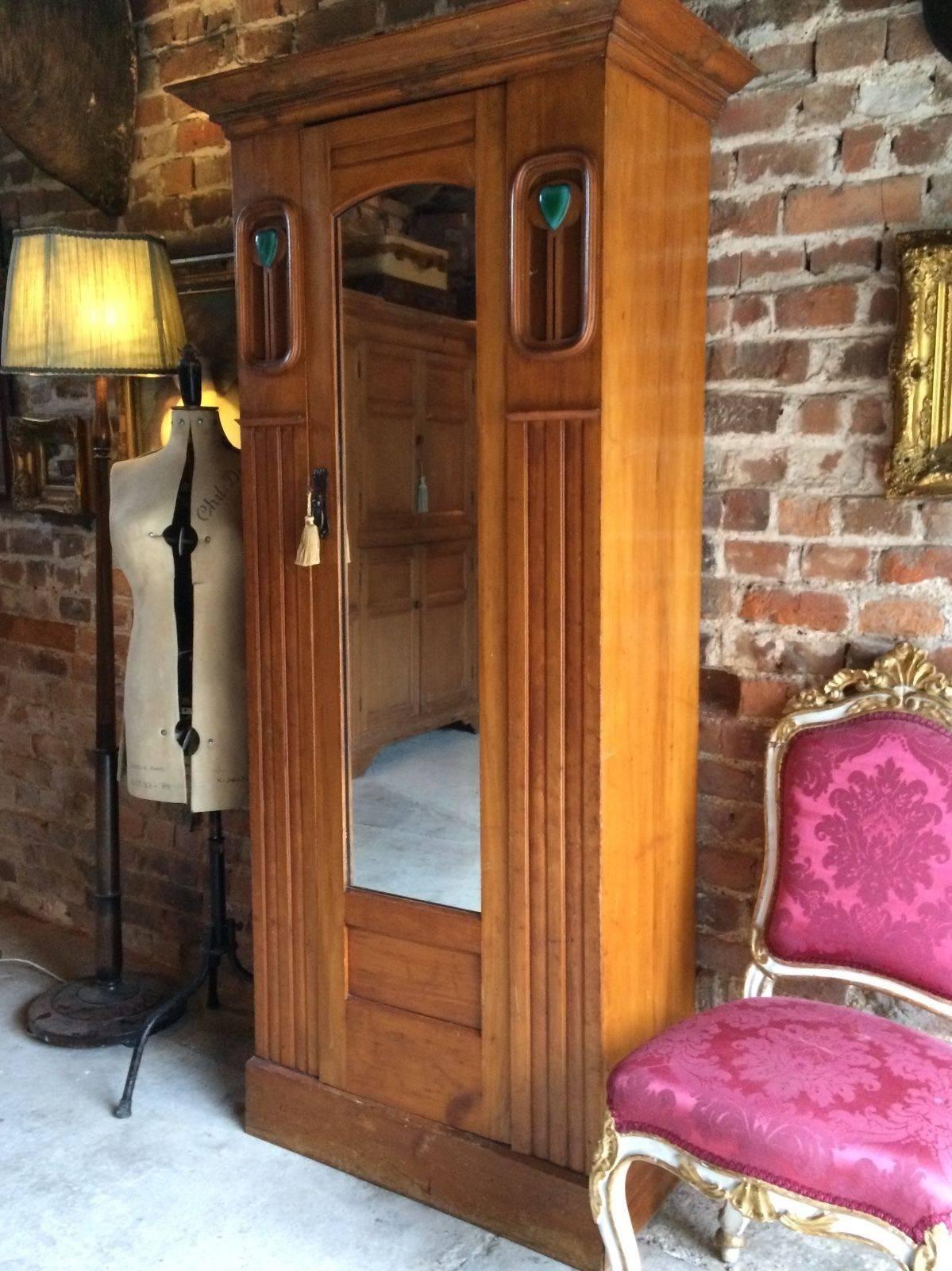 A beautiful Arts and Crafts walnut wardrobe with Ruskin style plaques, the moulded overhanging cornice above single panelled door with mirror to front enclosing brass hanging rail within, flanked by two panelled sections with turquoise heart shaped