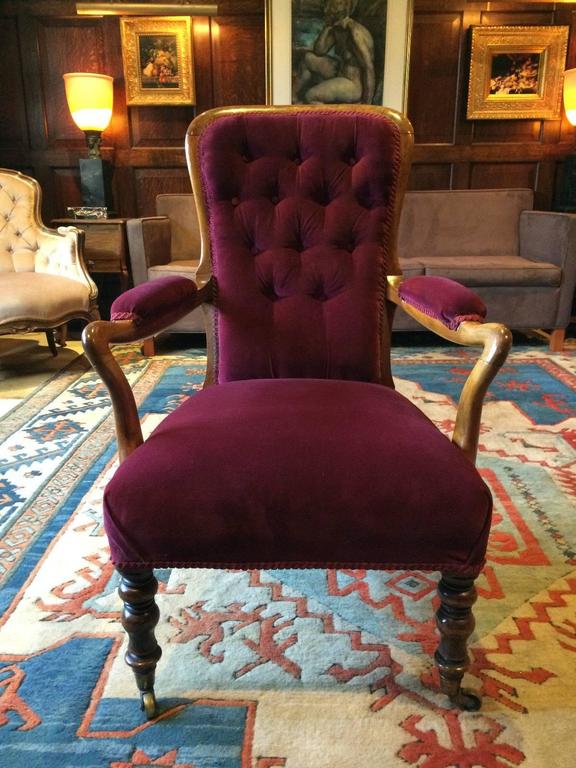 A beautiful late Victorian Mahogany framed open arm button-back salon chair with curved back and scrolling arms with pads, finished in a lush ‘Raspberry’ velvet material, standing on turned front legs and raised on brass casters, the armchair is