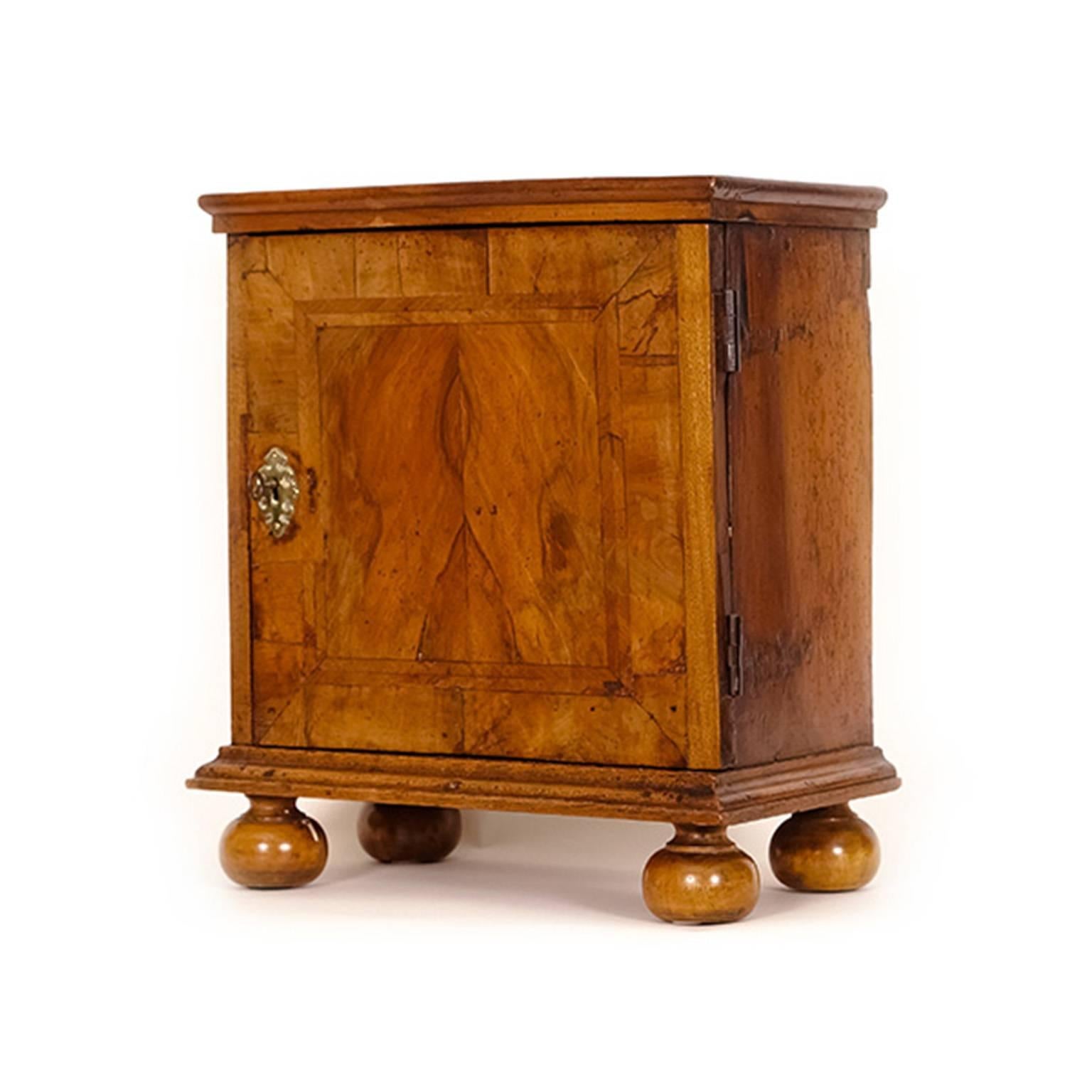 The door, inlaid with feather banding in a cross-banded border, opens to reveal a fitted interior of small drawers with brass drop handles and molding to the divides. Stands on a molded base with bun feet.